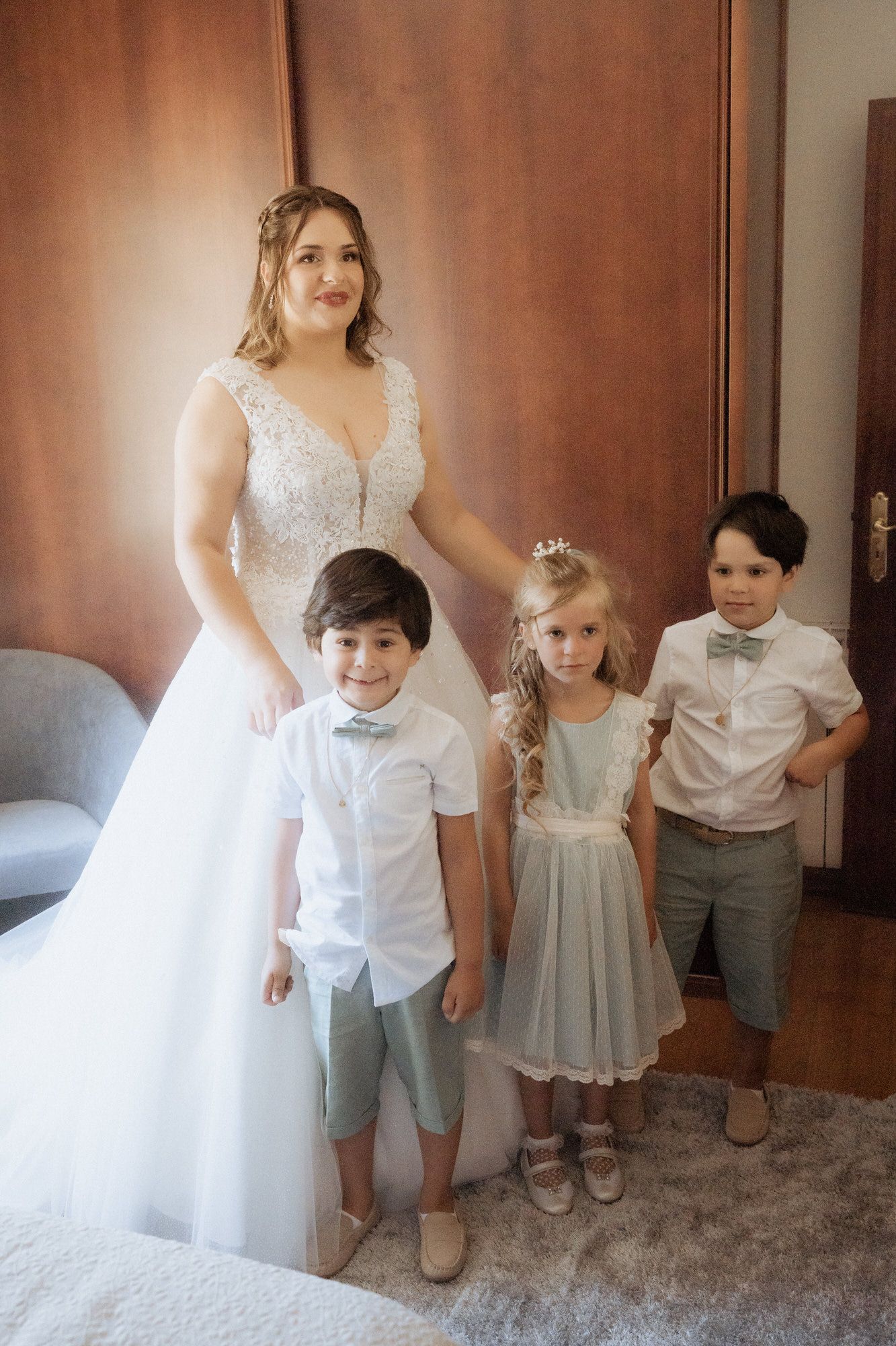 A bride in a white lace wedding dress poses with three young children in formal attire on a gray carpet.