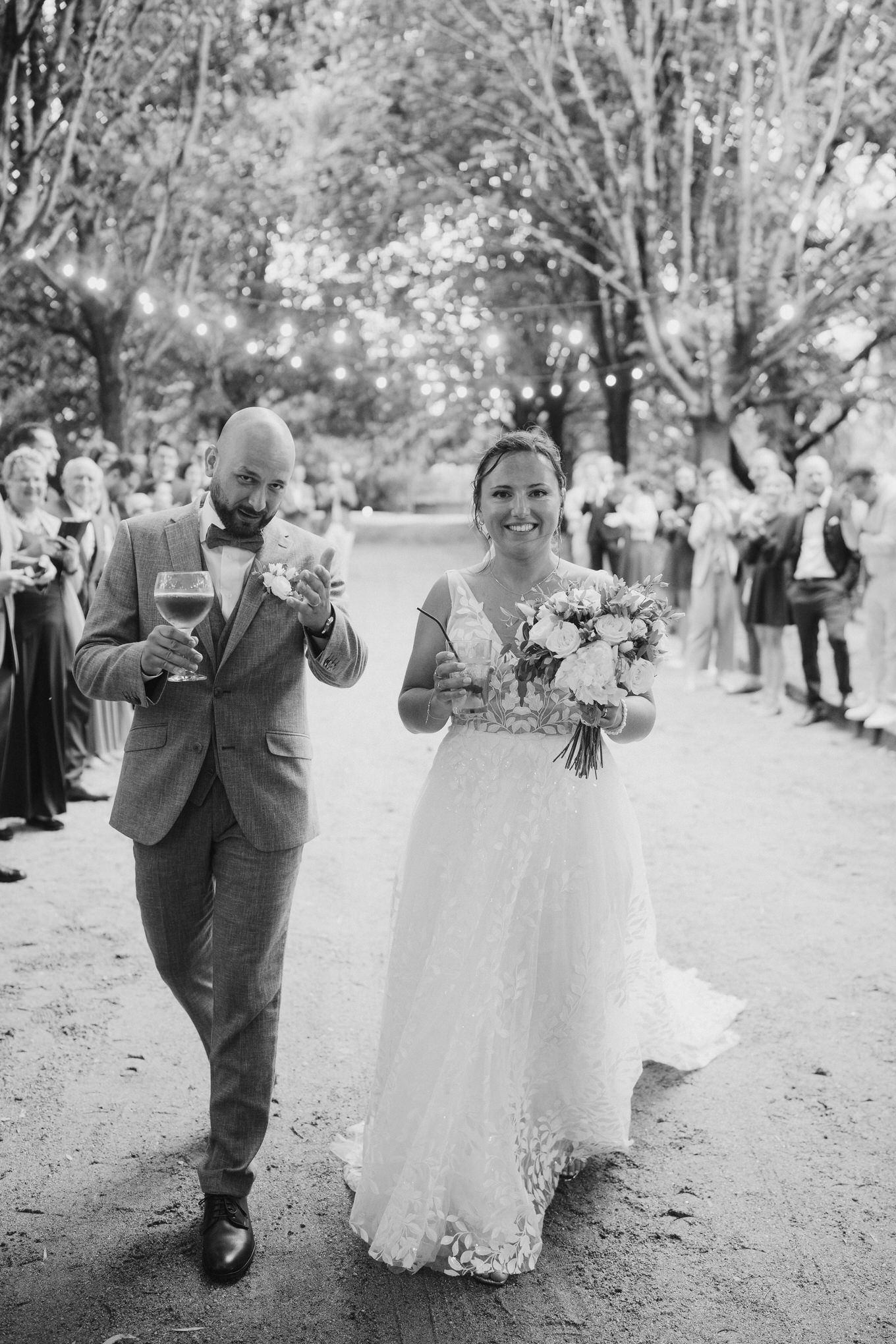 A joyful wedding couple walks down a tree-lined path while guests watch from the sides in black and white.