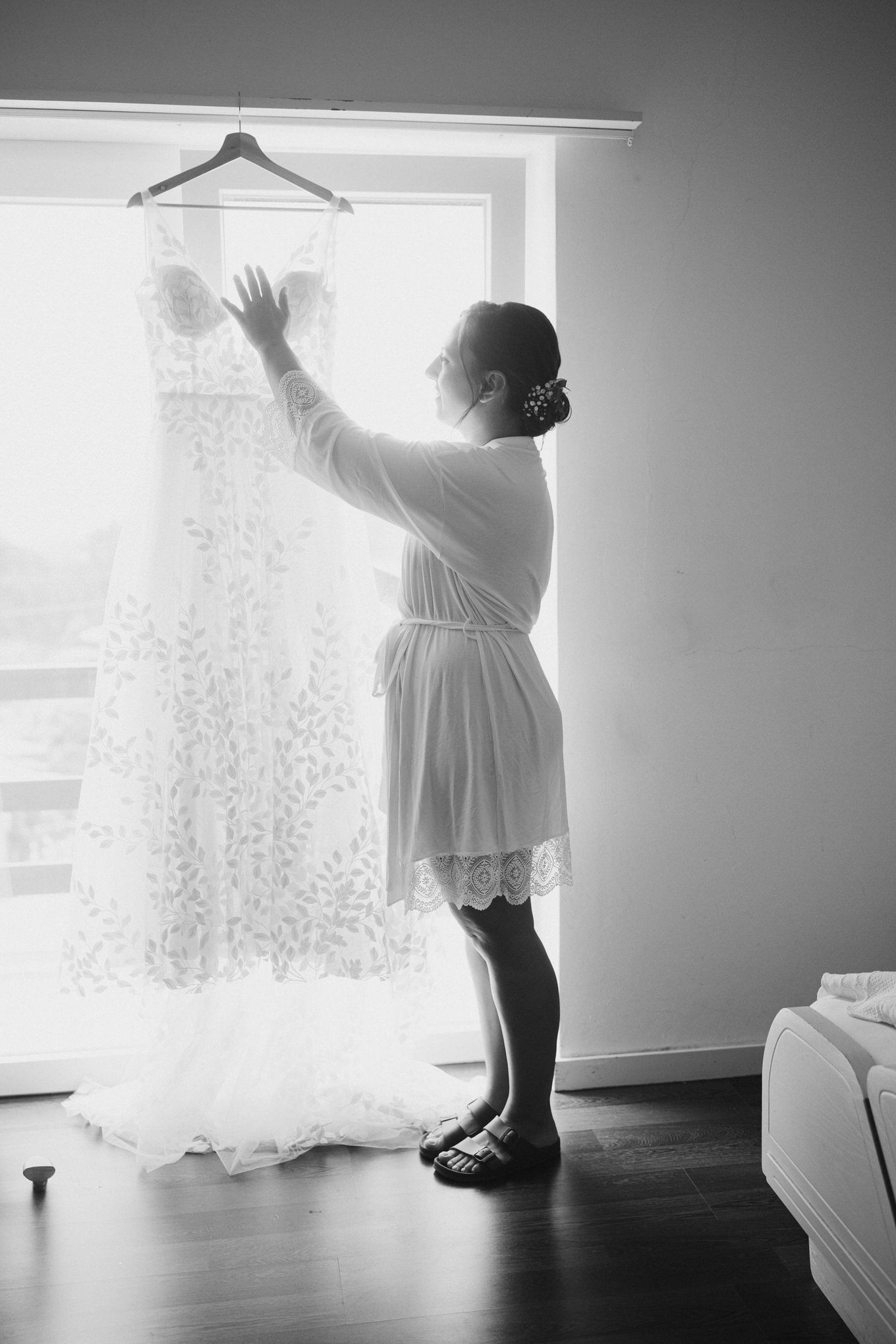 A person in a white robe reaches up to touch a wedding dress hanging near a bright window in a black and white photo.