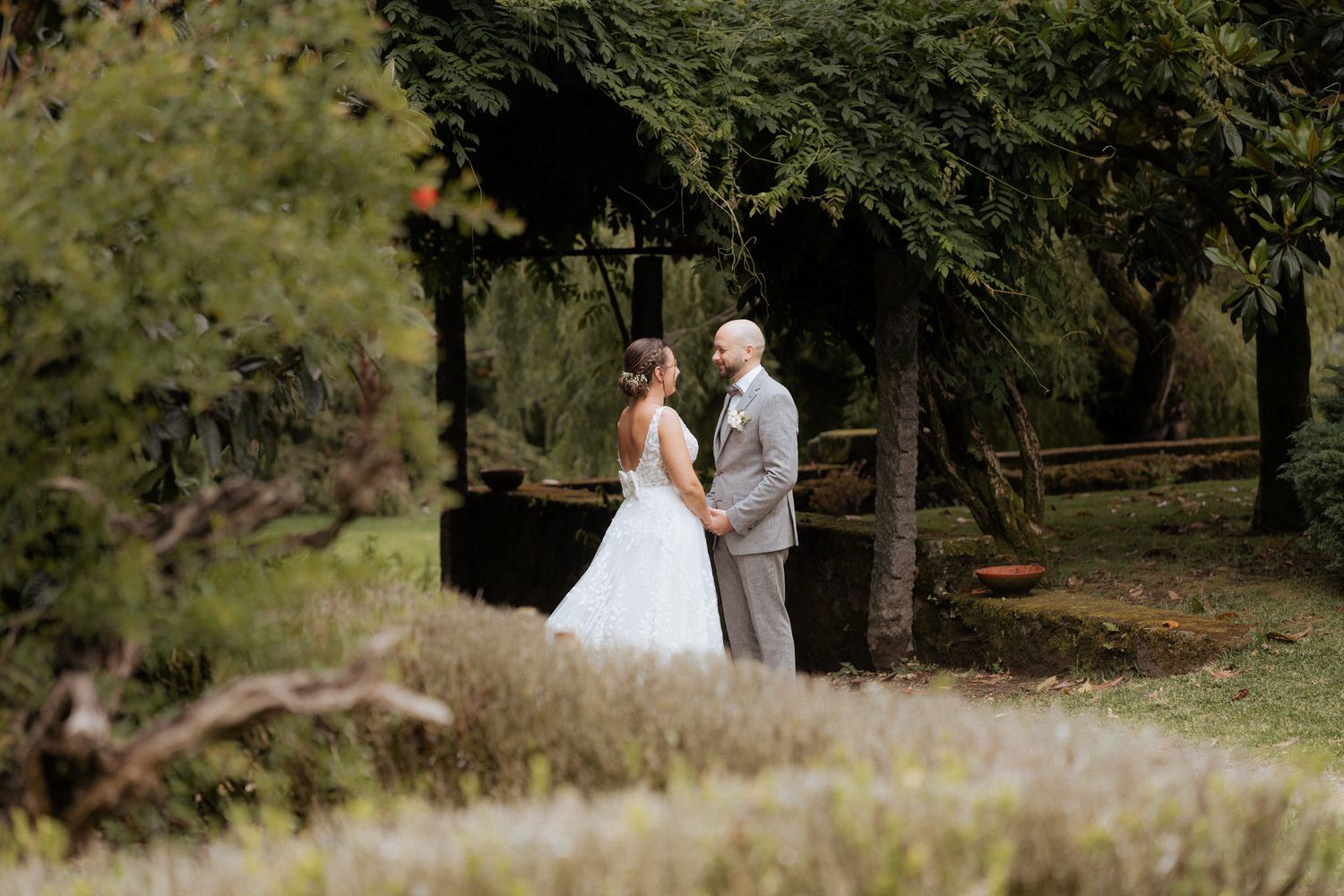 A couple shares an intimate moment outdoors in a wooded setting with natural greenery and soft light filtering through trees.