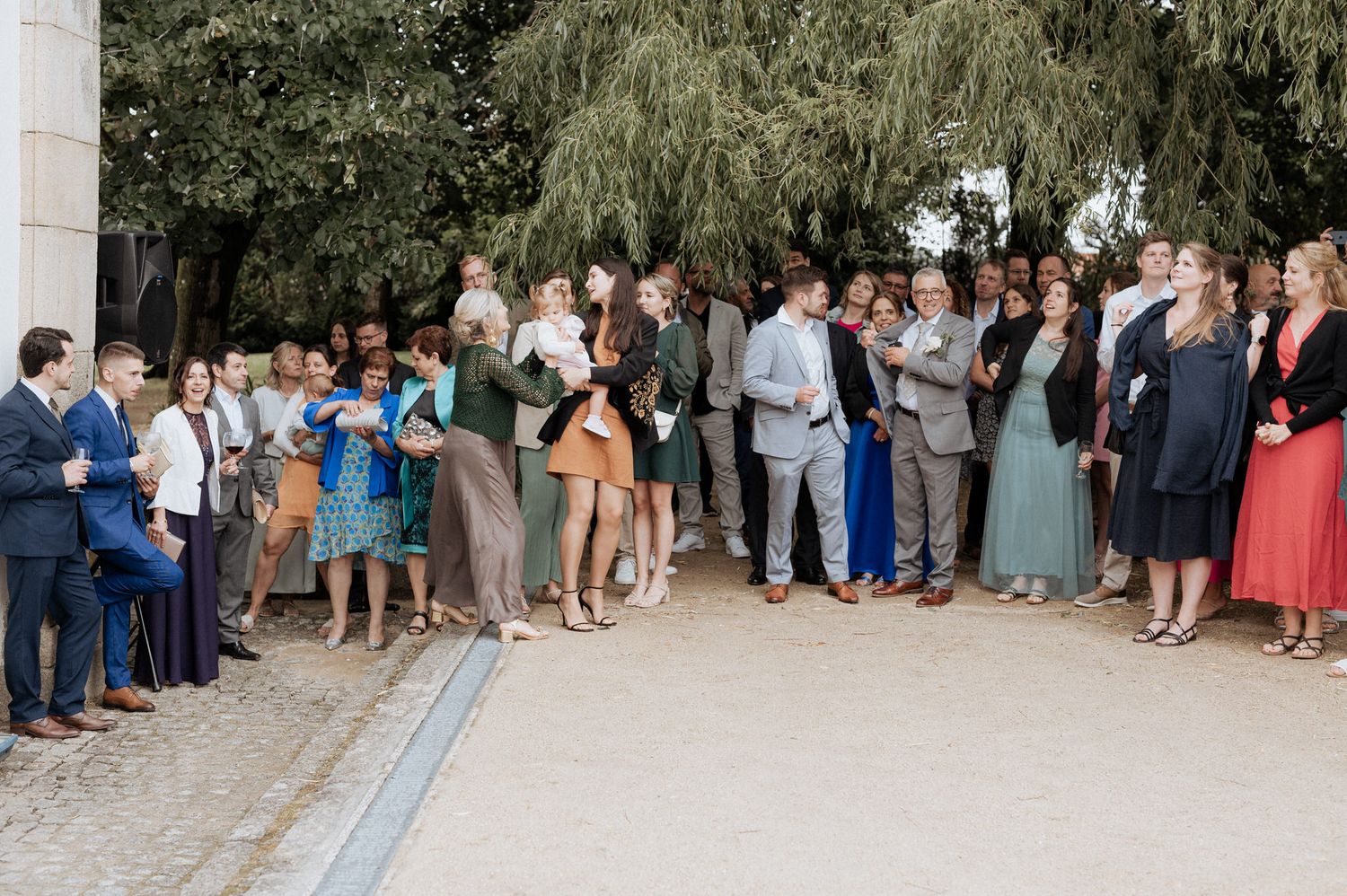 A large group of wedding guests gather outside on a stone pathway next to a willow tree.