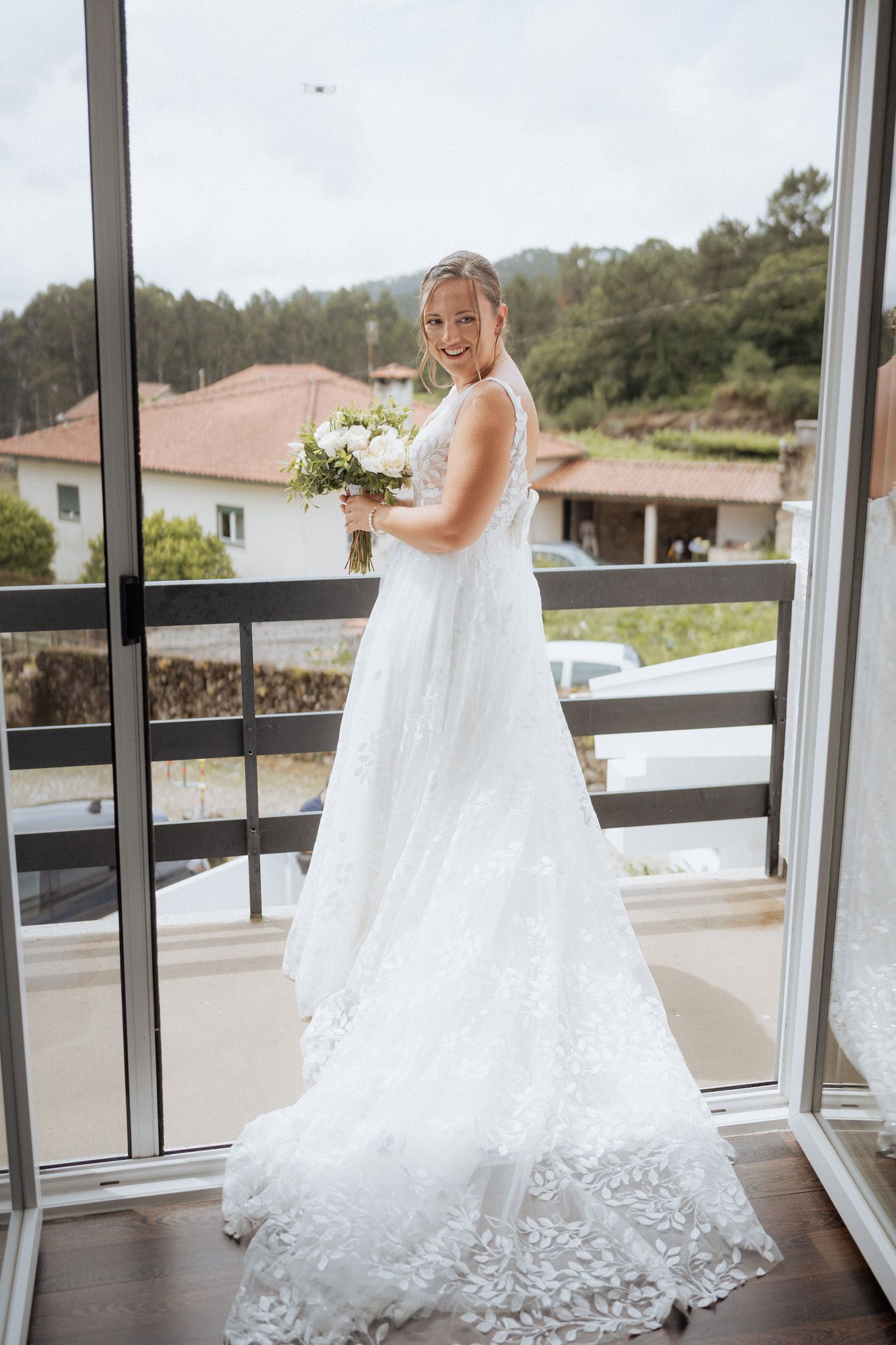 A bride in an elegant white lace wedding gown stands on a balcony holding a bouquet of flowers.