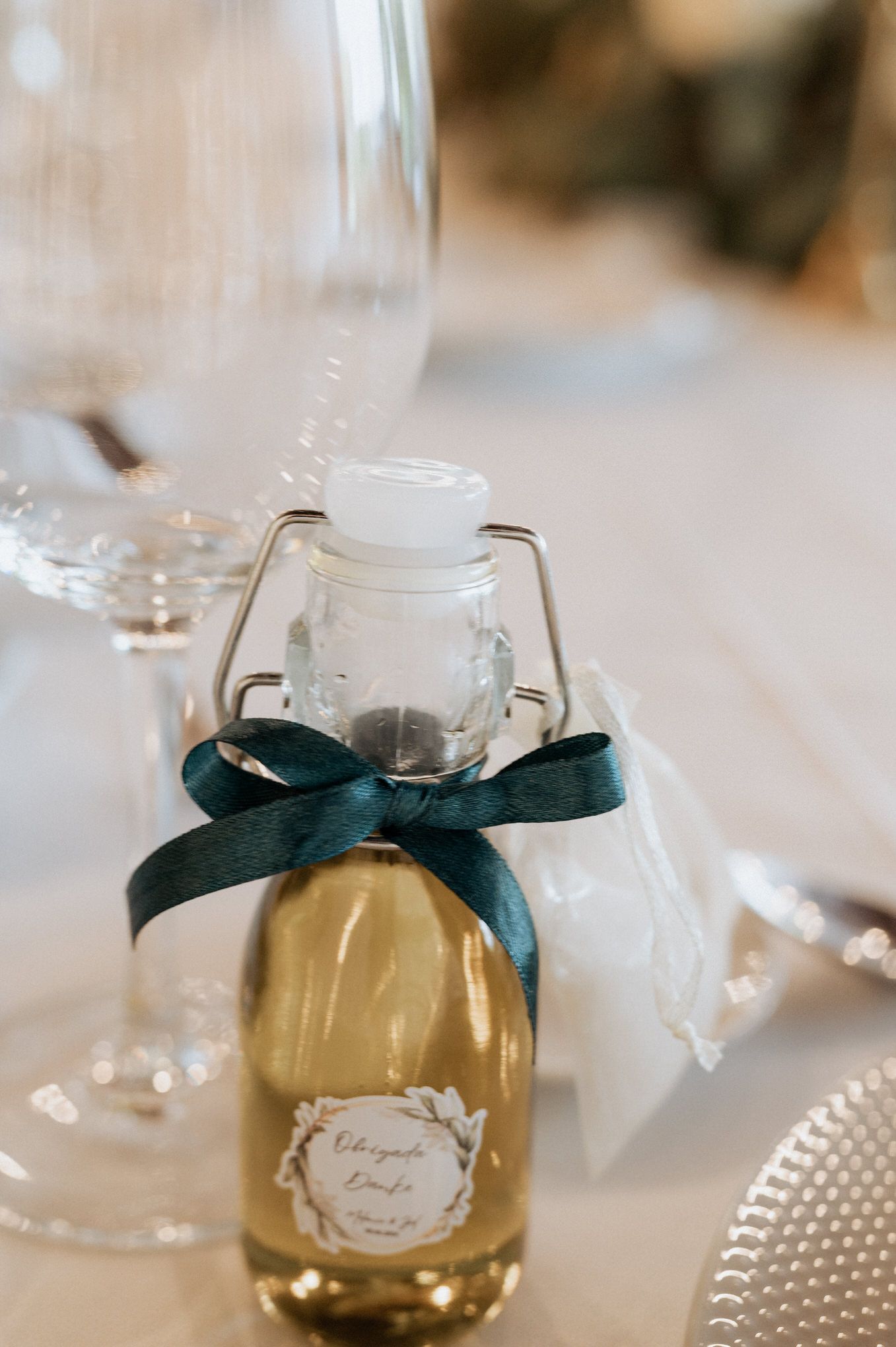 Small bottle of champagne favors with dark teal ribbon bow and custom label at an elegant wedding table setting.