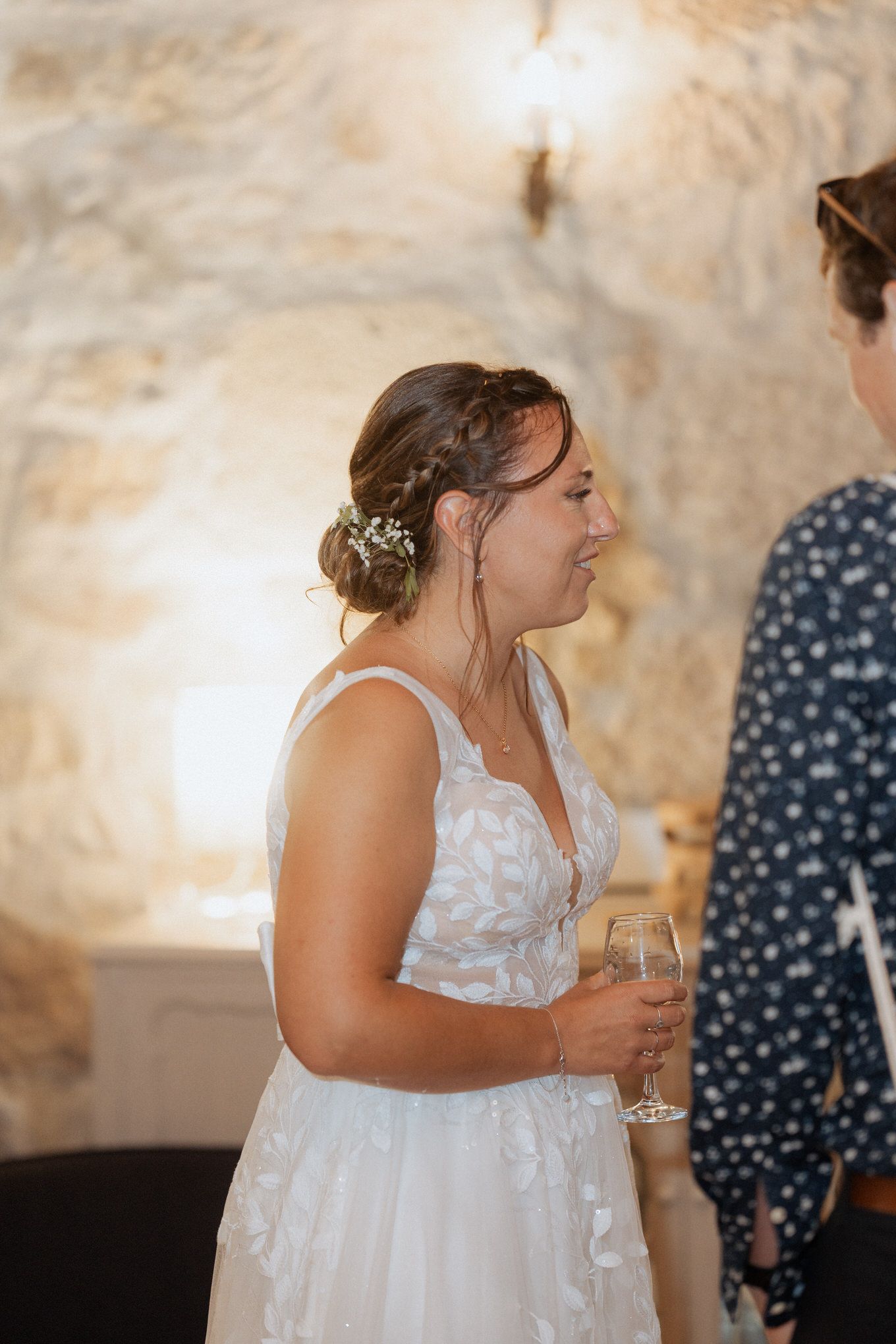 A person in a white lace wedding dress holds a champagne glass while smiling during an indoor celebration.