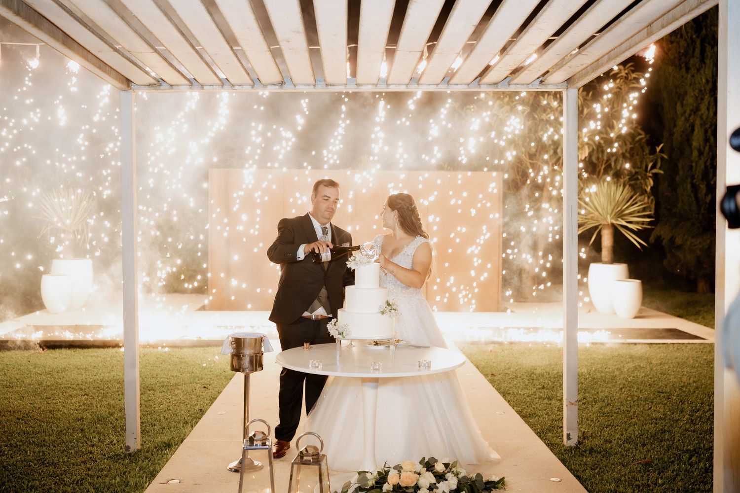 Couple celebrates their wedding under a white pergola with sparkling lights in an outdoor evening garden setting.