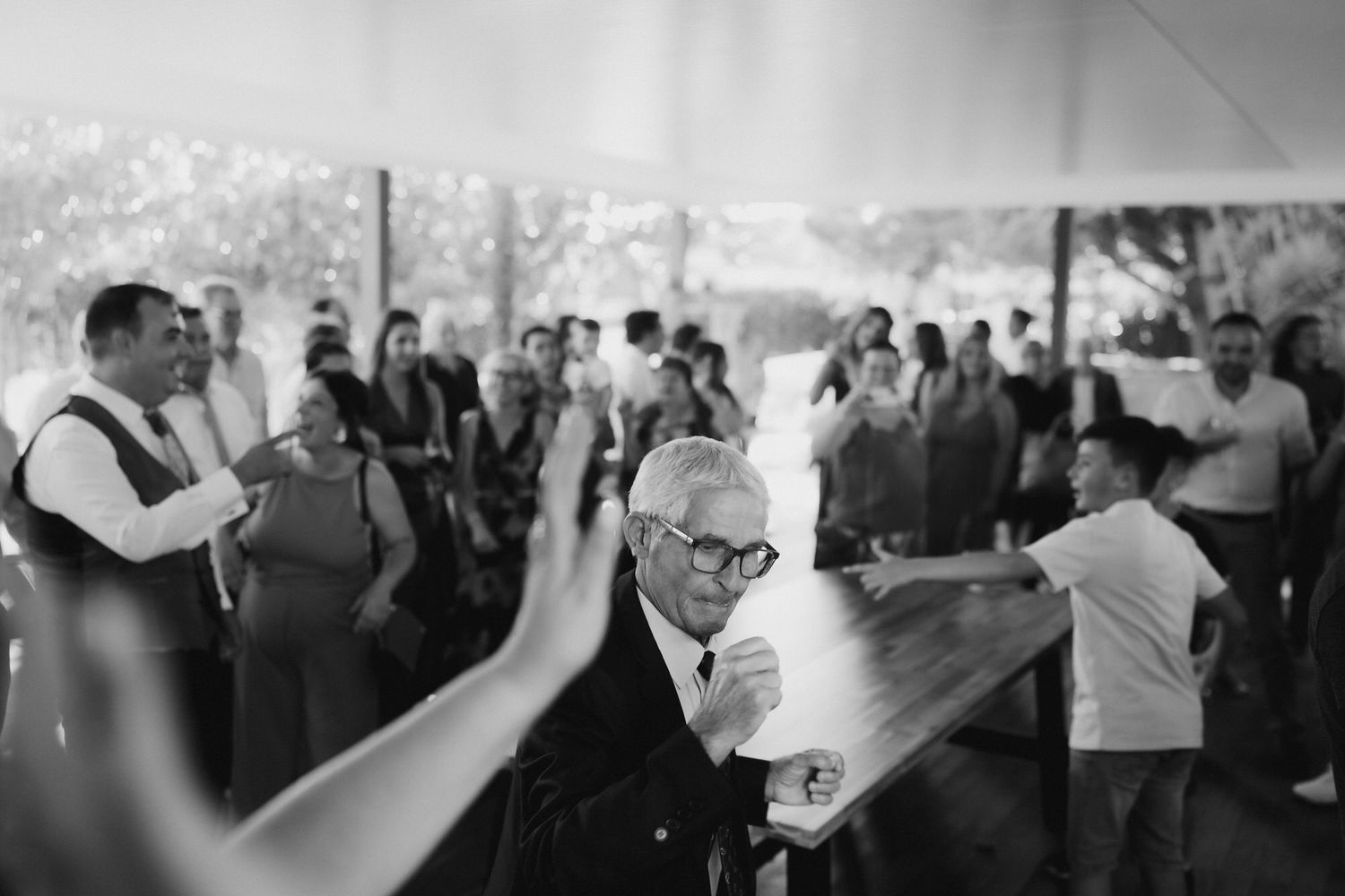 Wedding guests dance and celebrate together under a white tent during an outdoor reception celebration.