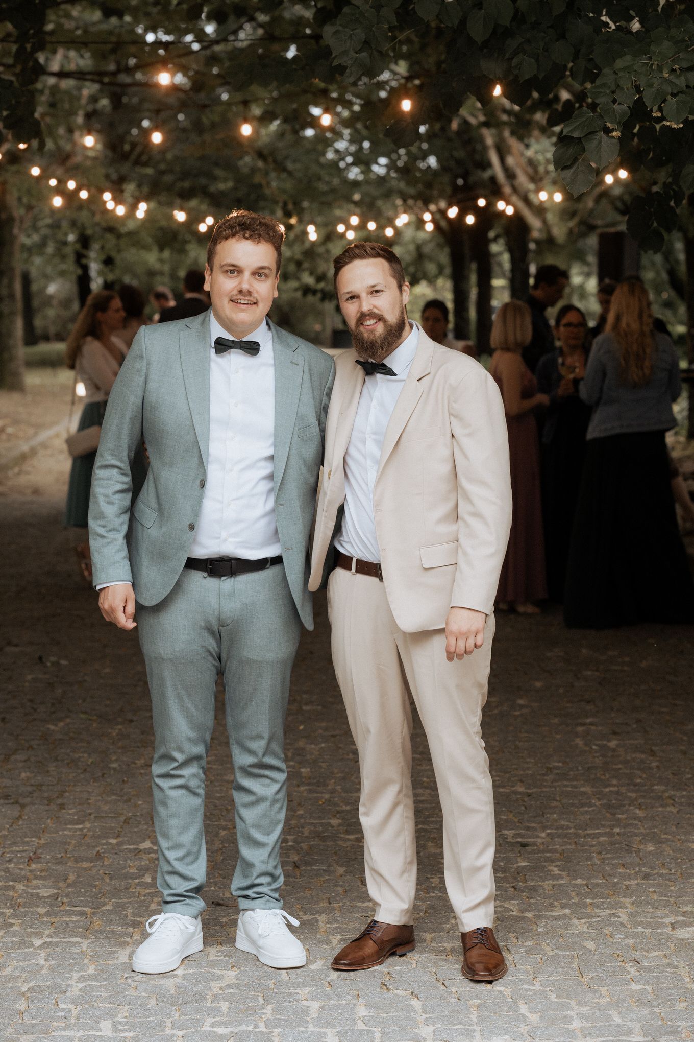 Two men in formal attire pose at an outdoor evening event with string lights overhead.
