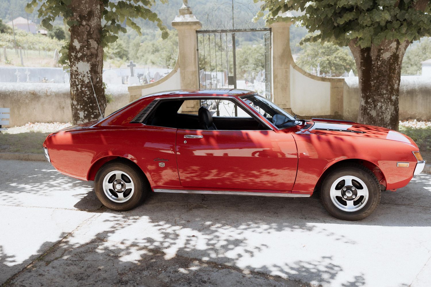 A classic red 1970s Mitsubishi car parked in front of a concrete wall with palm trees casting shadows.