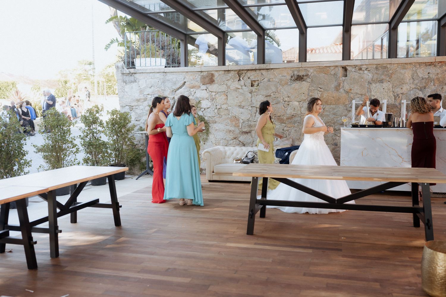 Wedding guests gather in a covered outdoor venue with wooden benches and stone walls during a reception.
