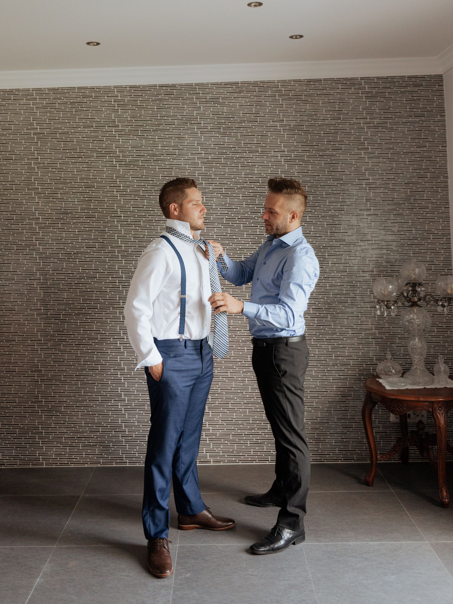 Two people in formal attire with suspenders and dress shirts adjusting each other's clothing against a brick wall.