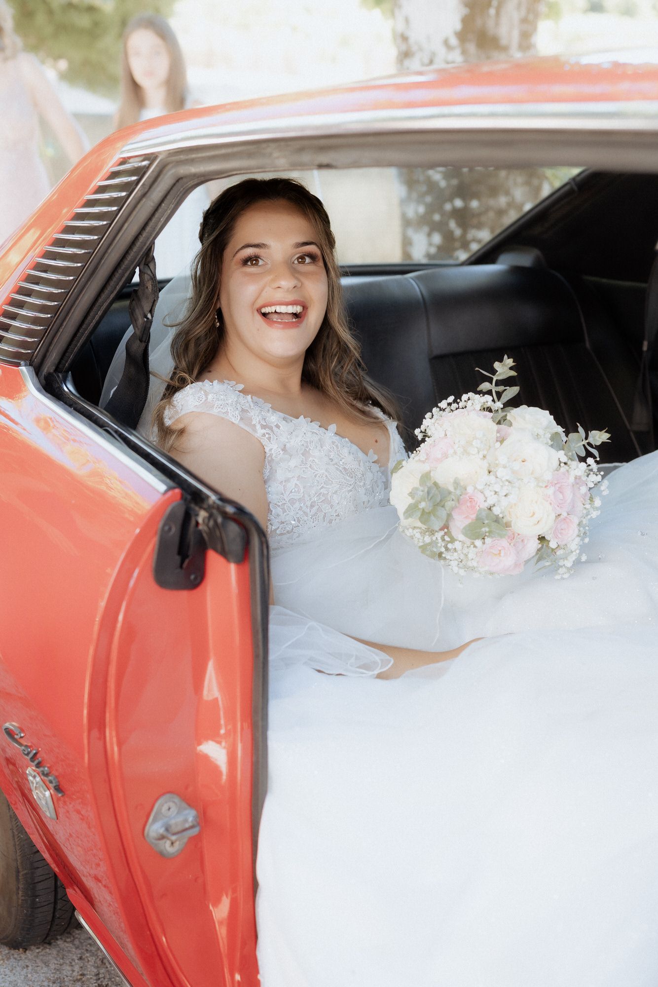 A bride in a white lace dress sits in a coral-colored vintage car while holding a soft pink and white floral bouquet.