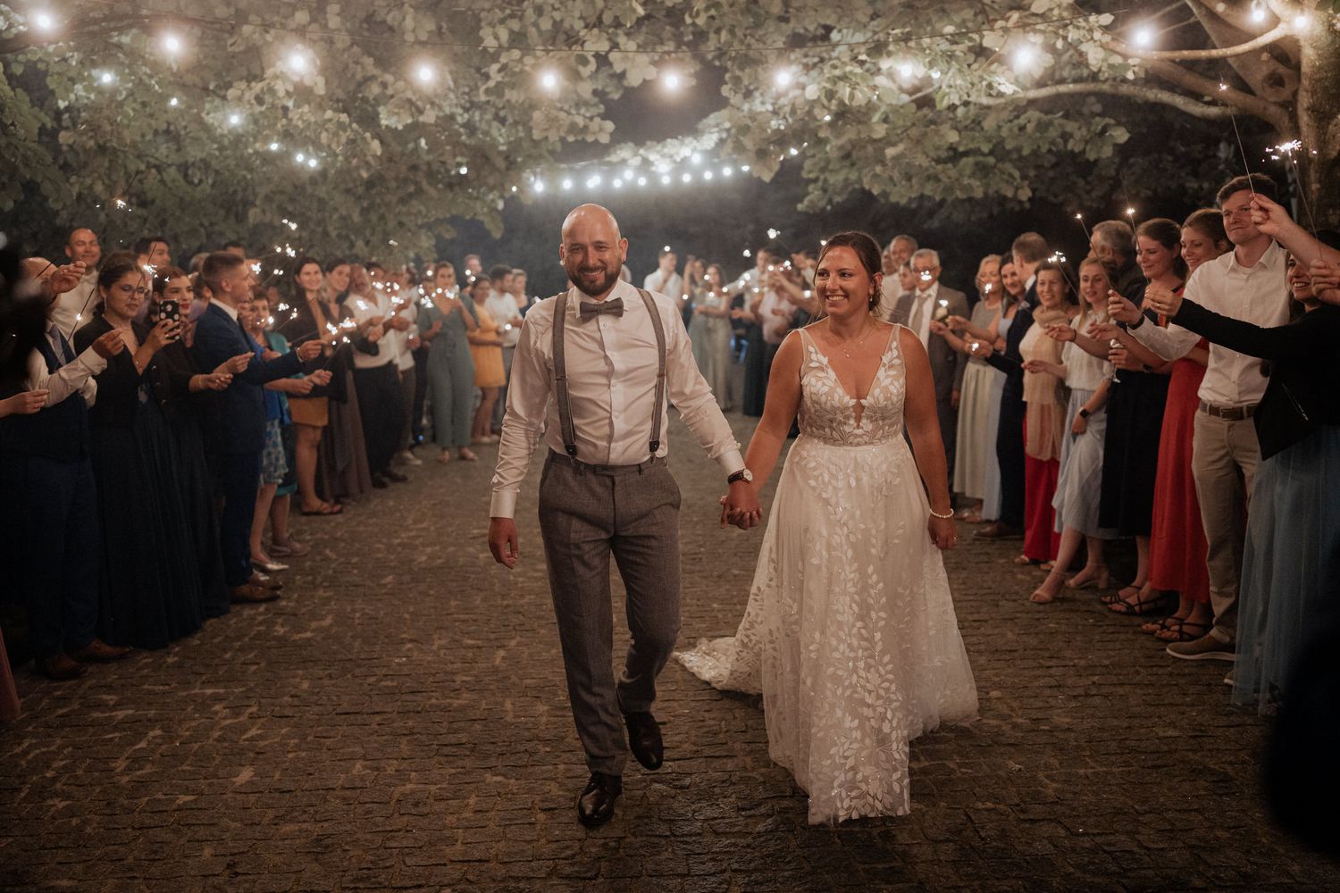 Newlyweds walk through sparkler exit line at outdoor evening reception as guests cheer and celebrate.