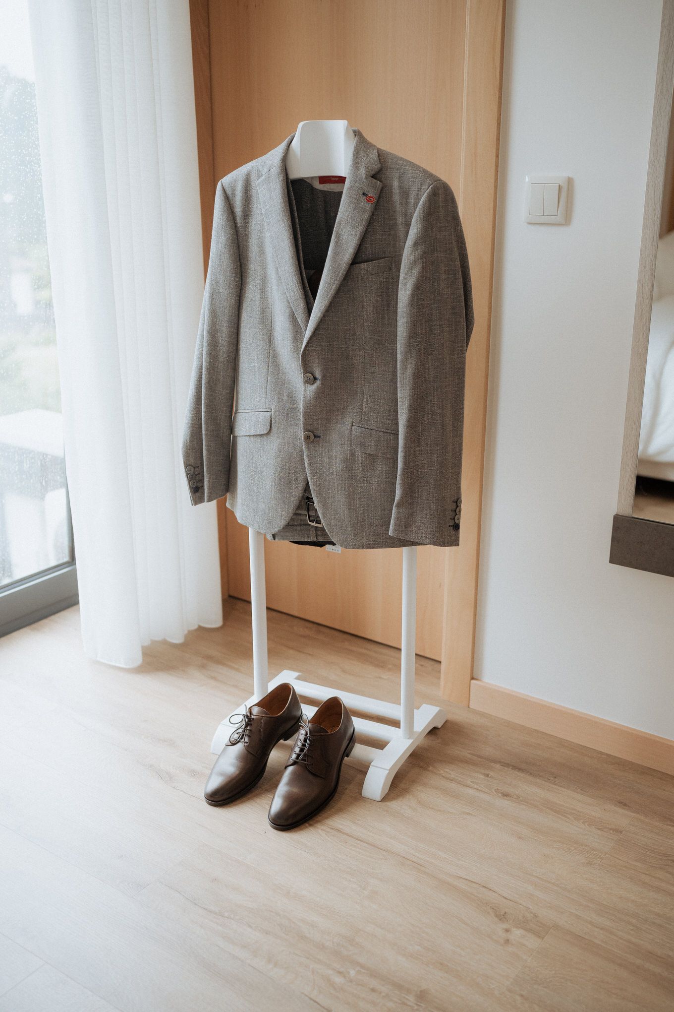 Grey suit jacket hanging on white stand with brown leather dress shoes beneath on wooden floor.