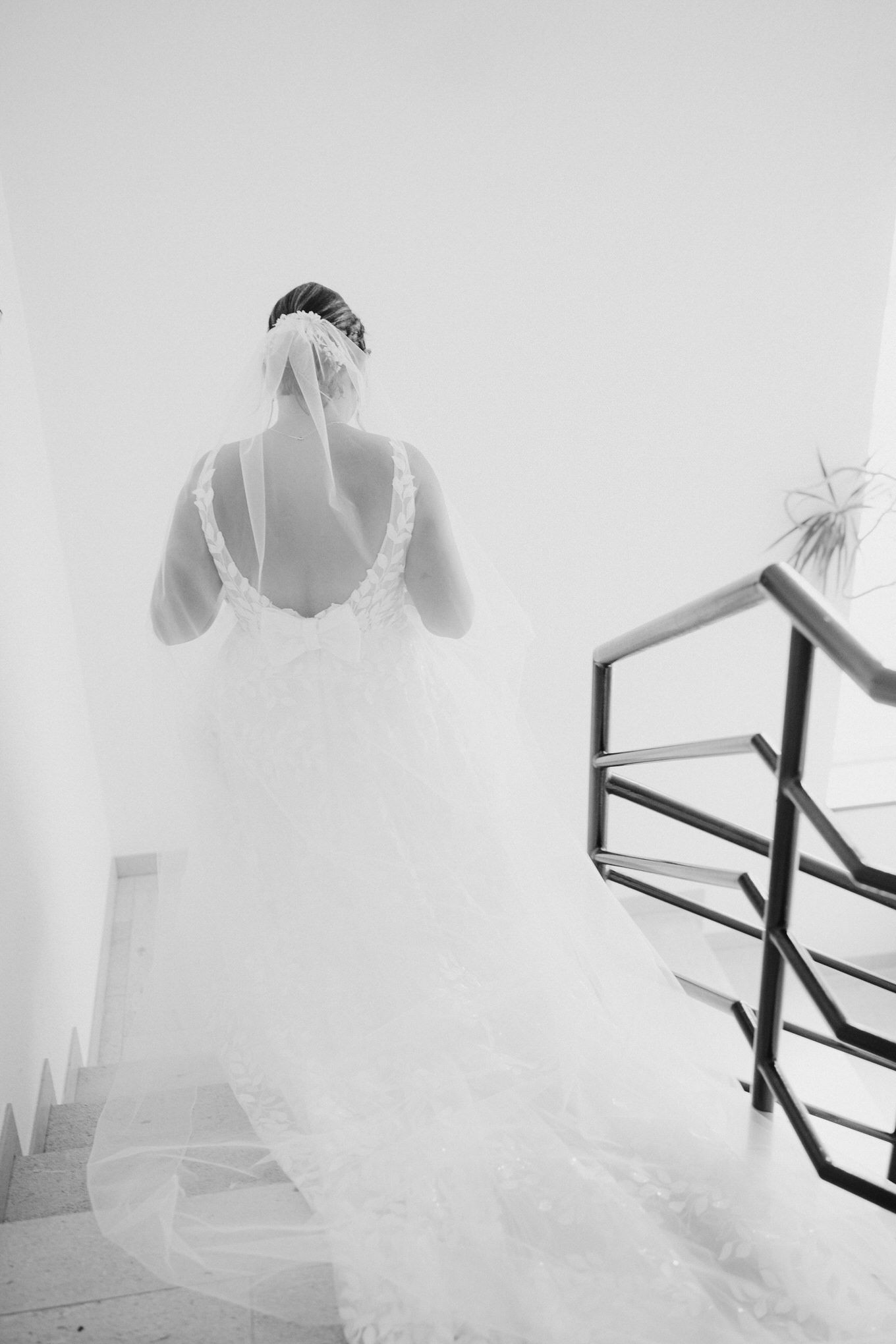 Dramatic black and white photo of a bride in flowing veil descending a staircase with modern railing.