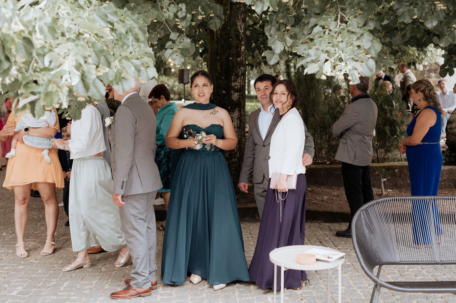 Guests in formal attire gather under a tree canopy at an outdoor event, with someone wearing an elegant teal gown.