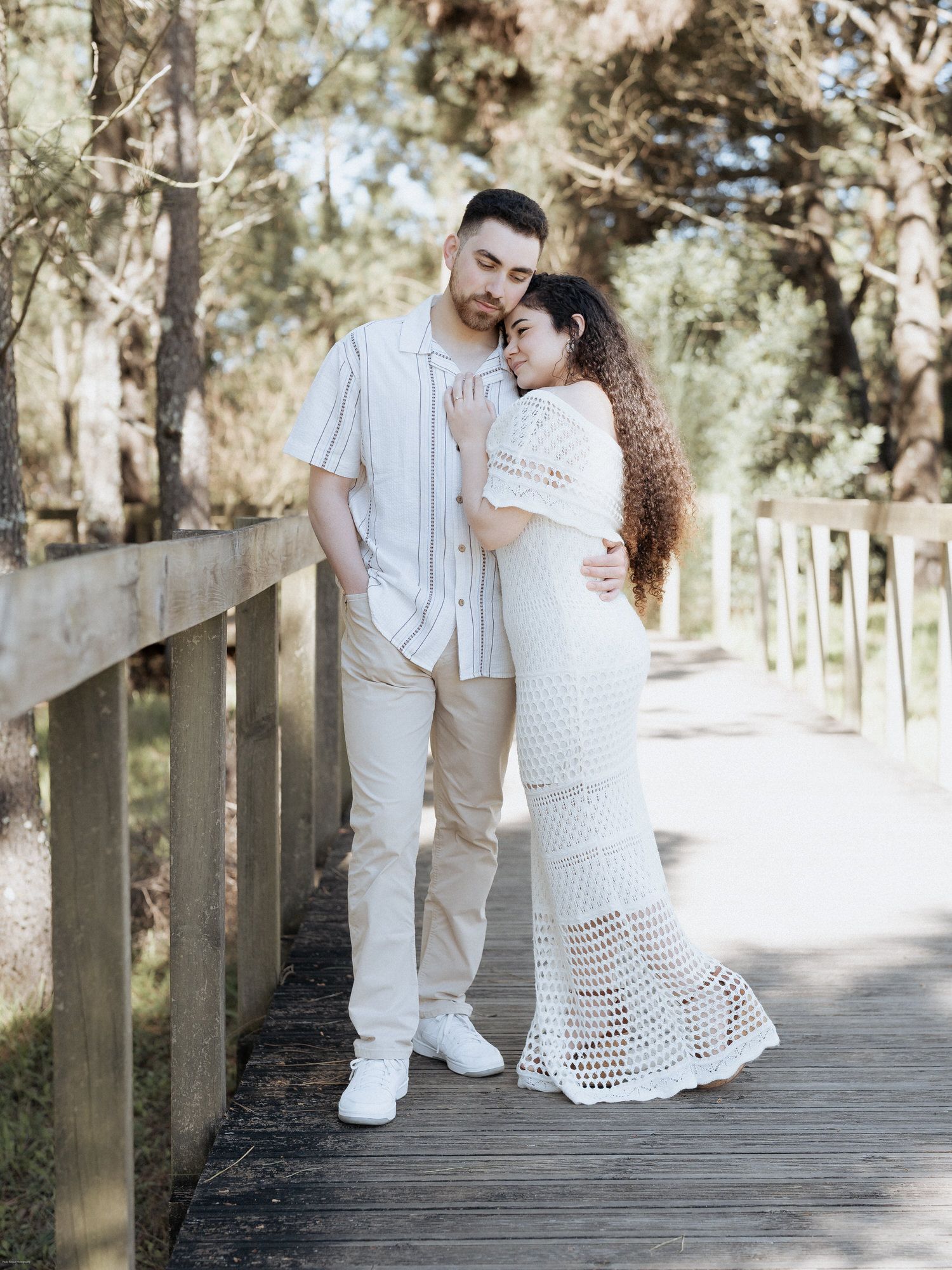 Um casal com elegantes trajes brancos abraça-se numa passarela de madeira rodeada por pinheiros num dia ensolarado.