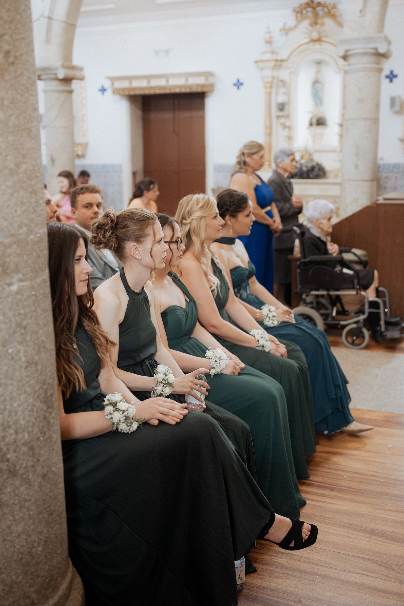 Wedding party attendants in dark emerald green formal dresses sit in a row inside an ornate church sanctuary.