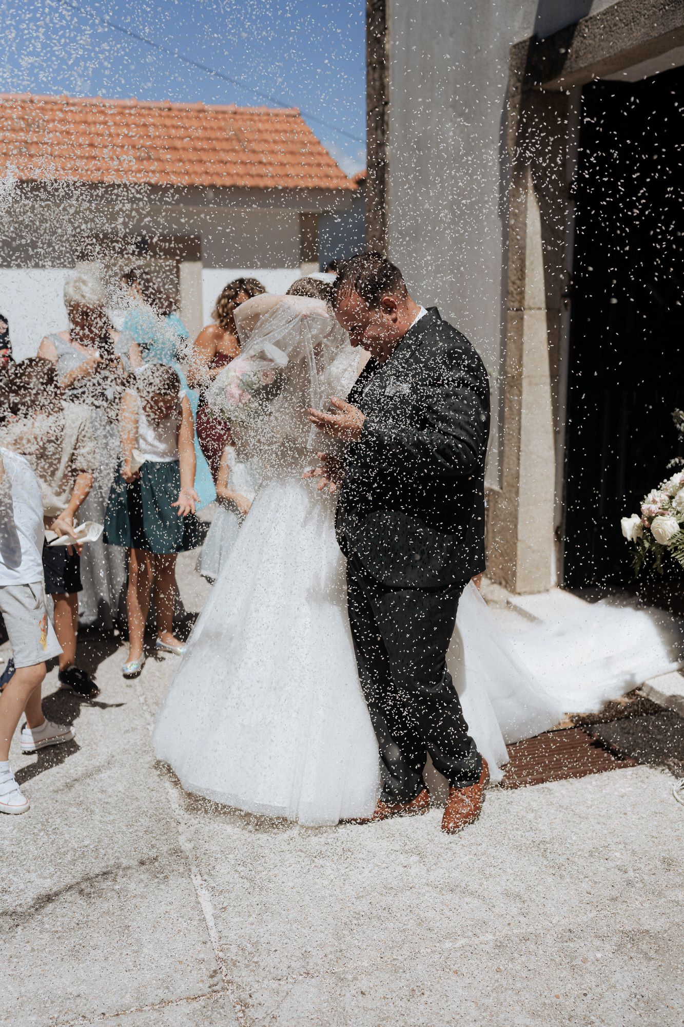 A couple shares their first kiss as newlyweds while guests shower them with confetti outside a church entrance.
