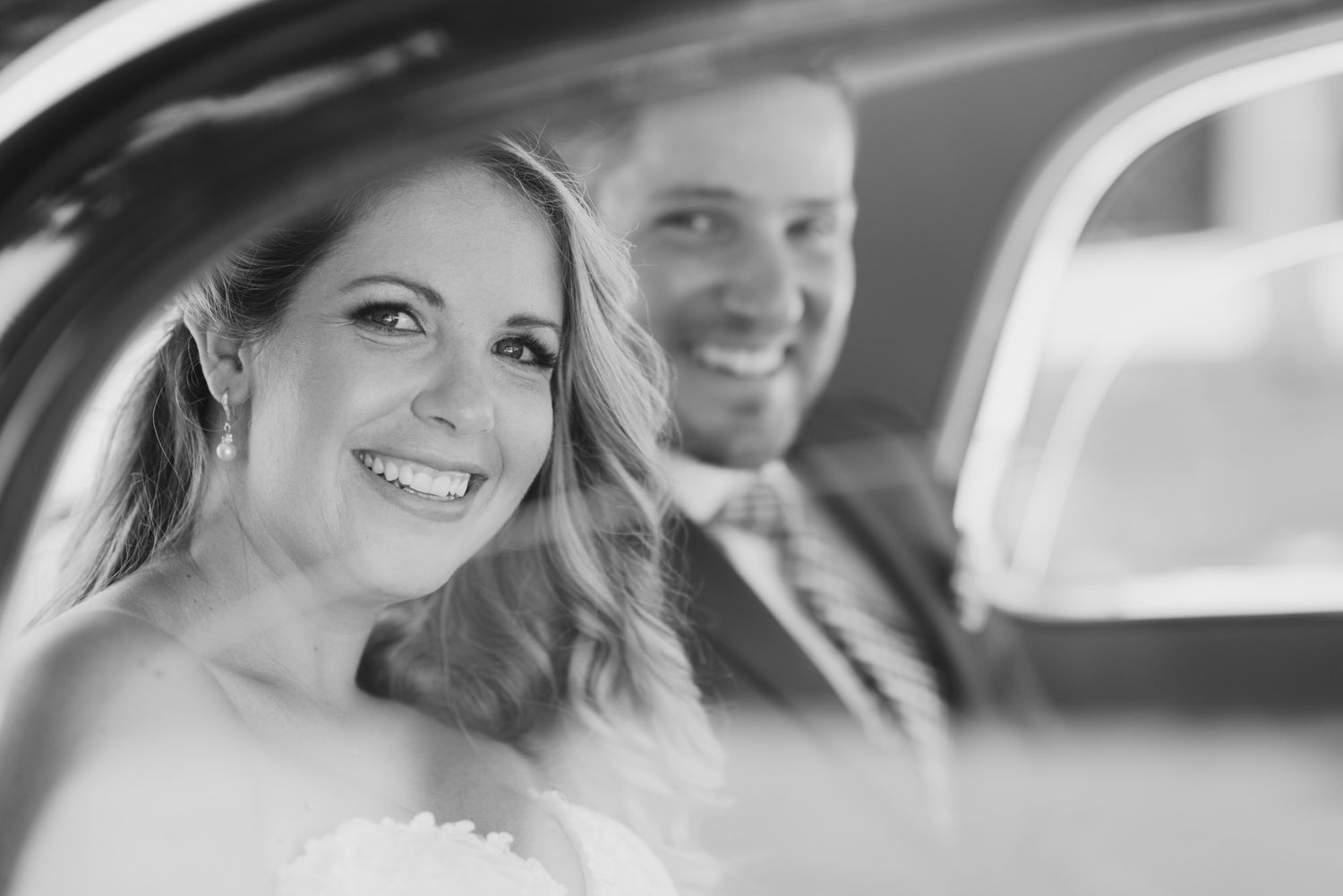 Happy newlyweds smiling together in back seat of wedding car, captured in black and white photograph.