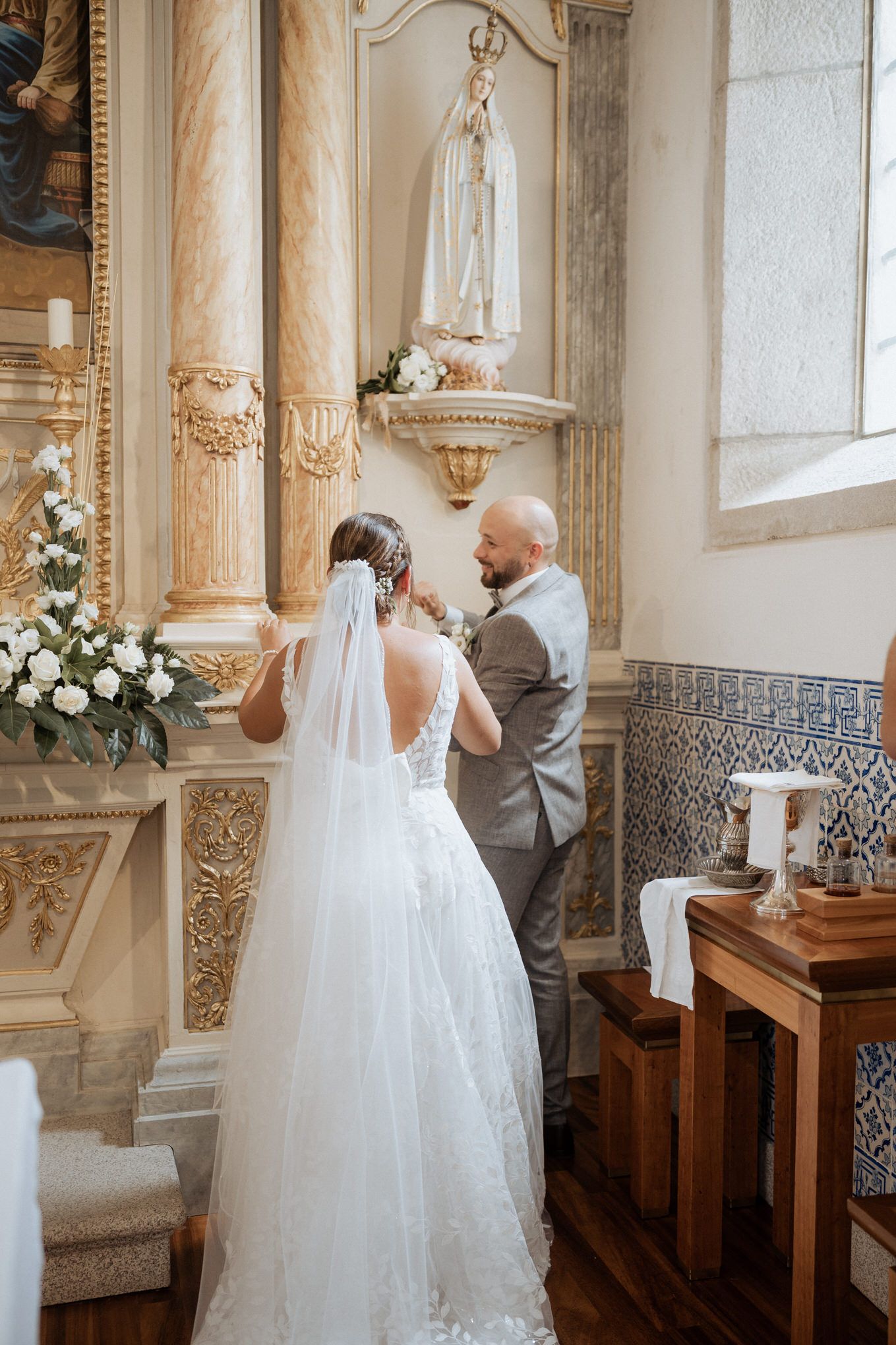 A bride in a lace wedding dress and groom in gray suit stand in an ornate church interior with religious statuary.