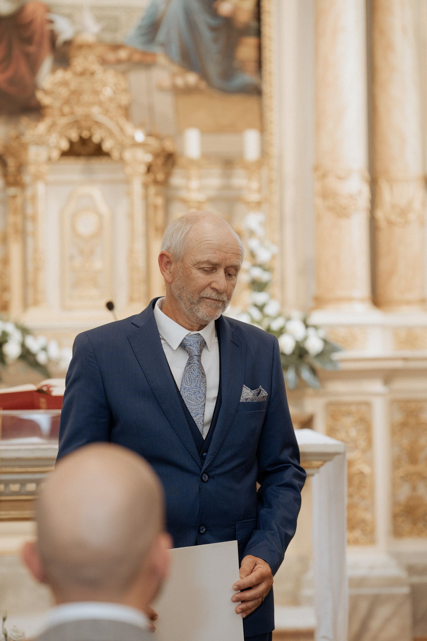 An older gentleman in a navy suit and tie stands at a church altar surrounded by ornate baroque decorations.