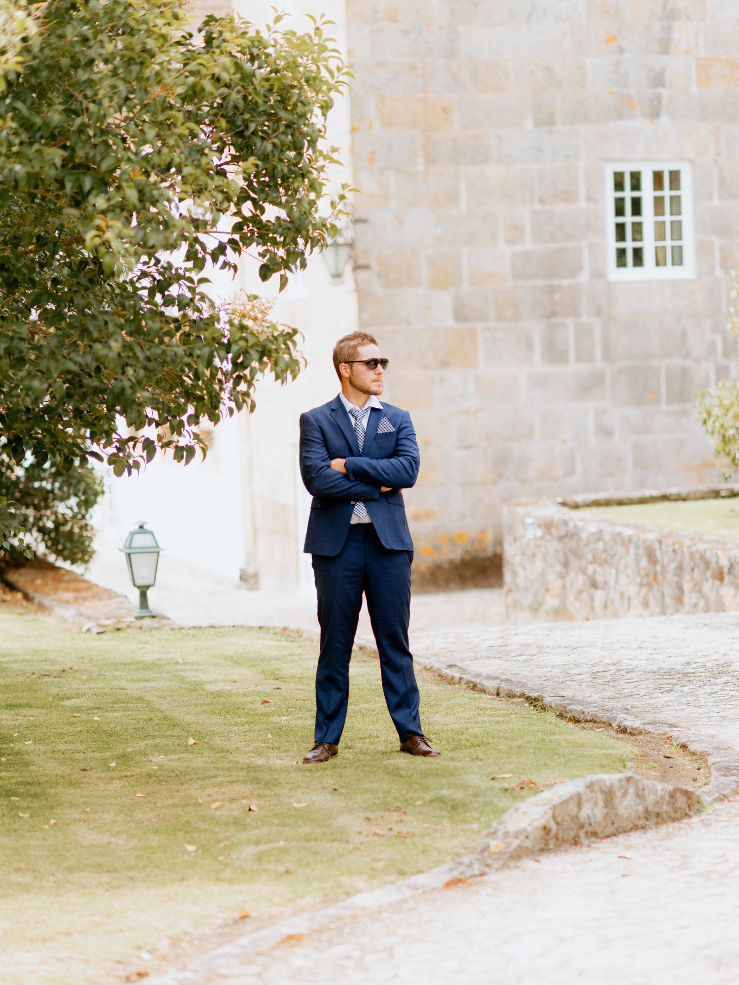 A person in a navy blue suit and sunglasses stands confidently with crossed arms on manicured grounds near stone walls.