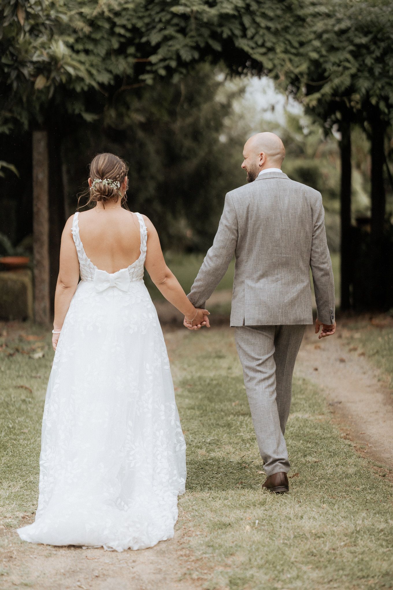 Couple walking hand in hand on garden path, with bride in backless lace gown and groom in light gray suit.