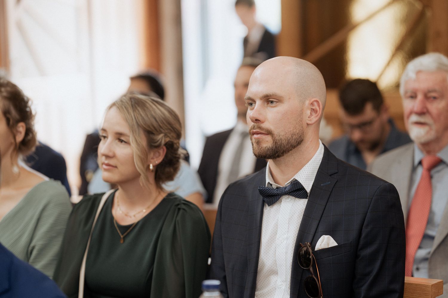 Wedding guests sitting quietly during a ceremony, with a person in a black suit wearing a bowtie.