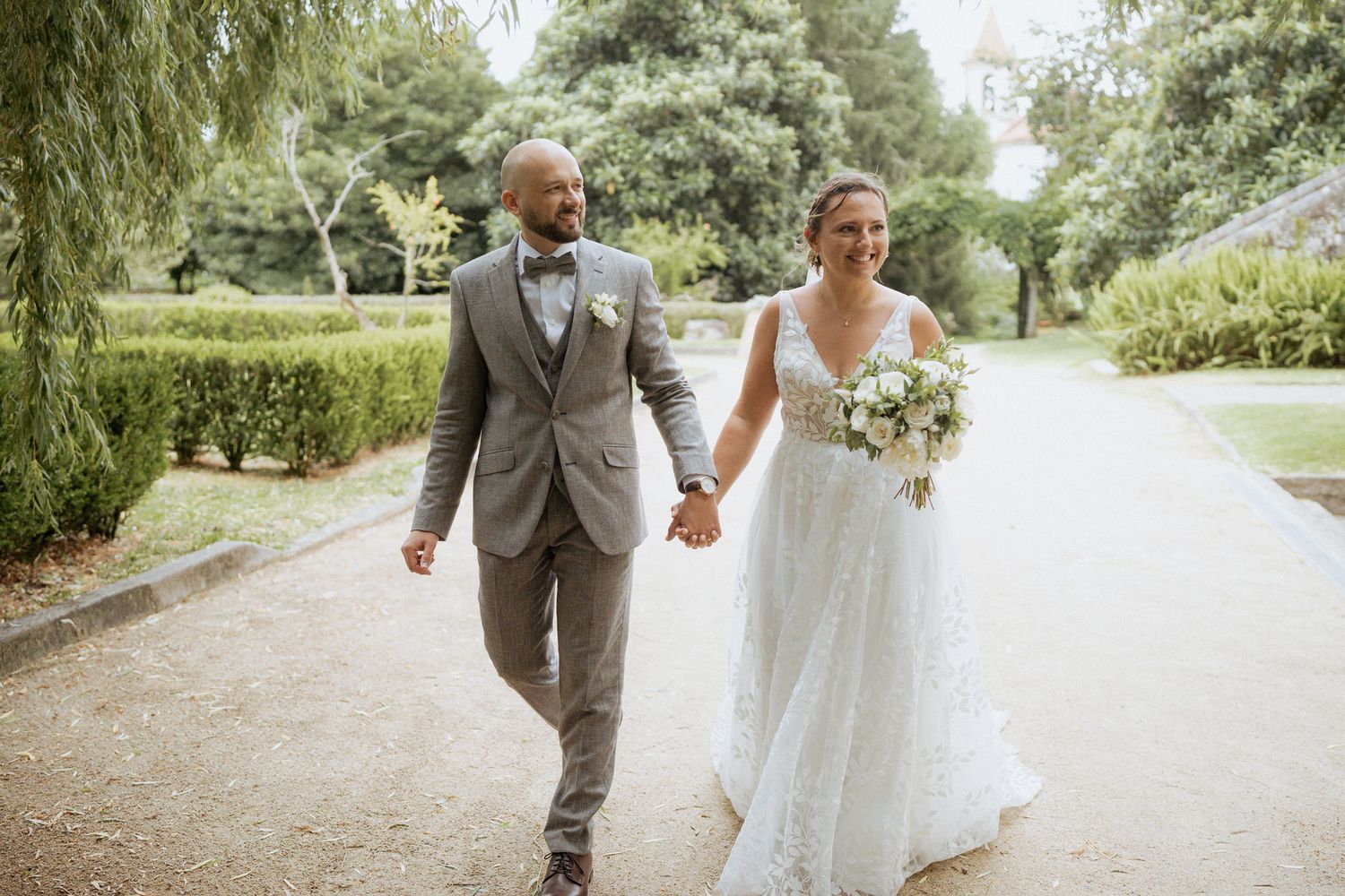 A happy couple walks hand in hand down a garden path on their wedding day, surrounded by lush greenery.