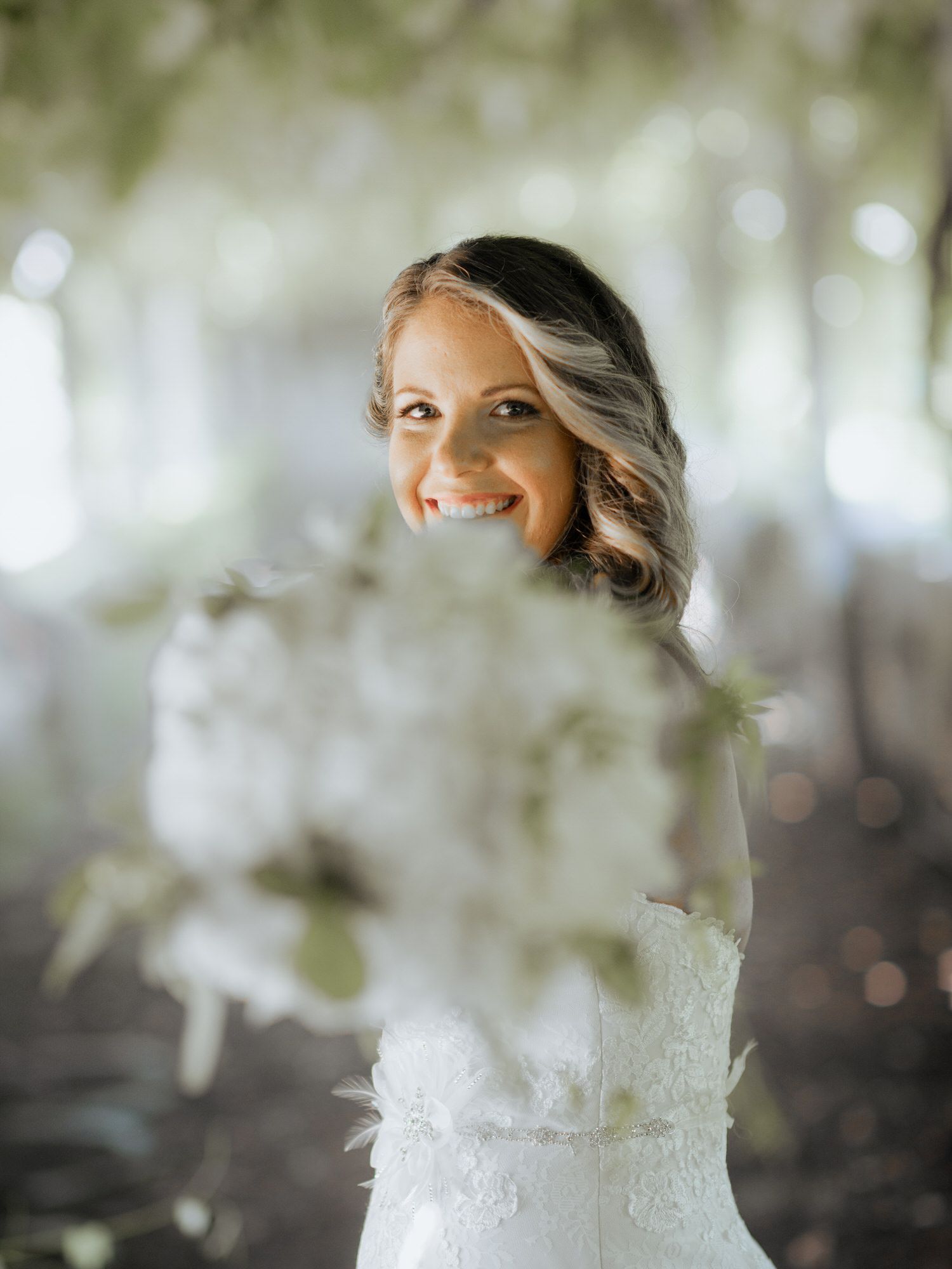 A radiant bride in a white lace dress smiles through delicate spring blossoms in a dreamy outdoor setting.
