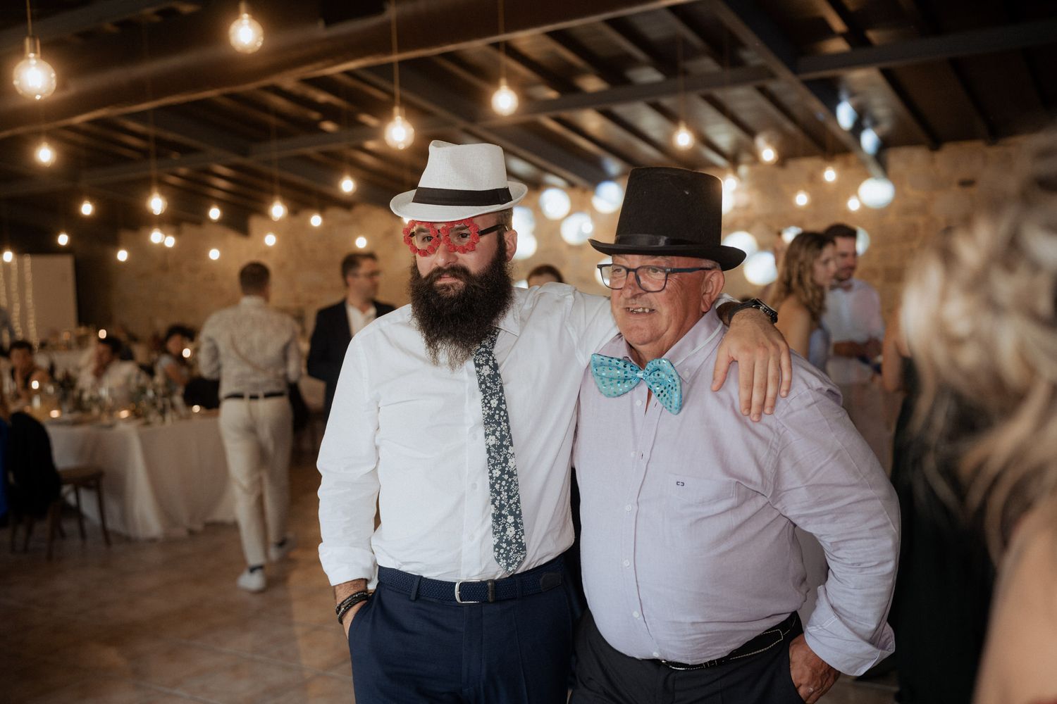 Two guests in festive attire share a warm moment at an indoor reception with string lights overhead.