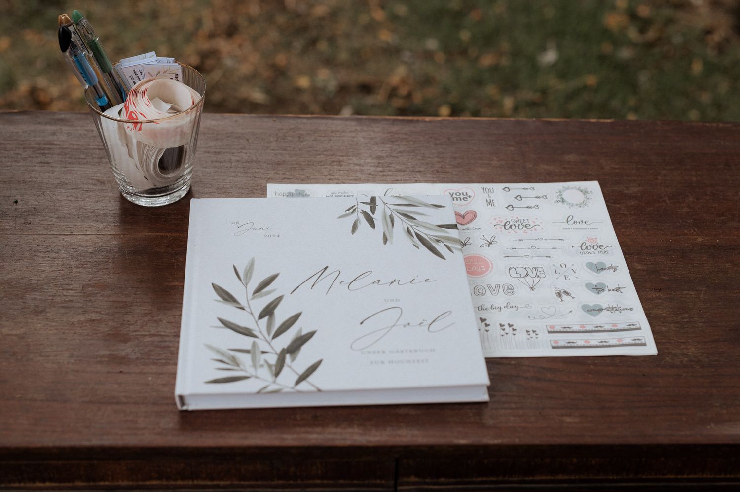Wedding invitation cards with olive branch designs and a milkshake drink sit on a rustic wooden table.