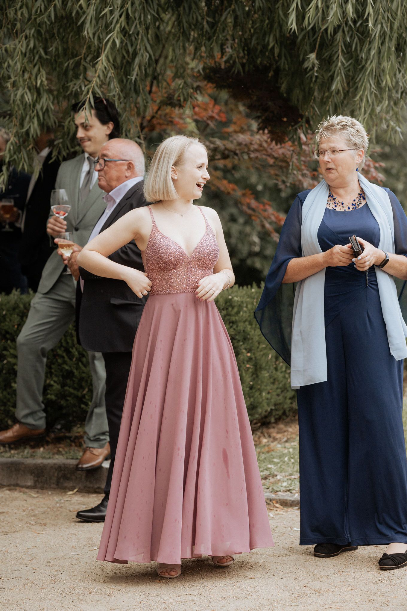 Wedding guests in formal attire gather outdoors under a willow tree during an elegant celebration.