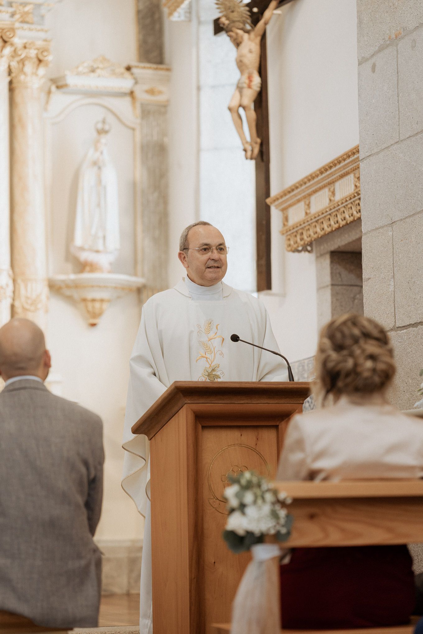 A Catholic priest in white vestments stands at a wooden lectern during a church ceremony with religious decor in background.