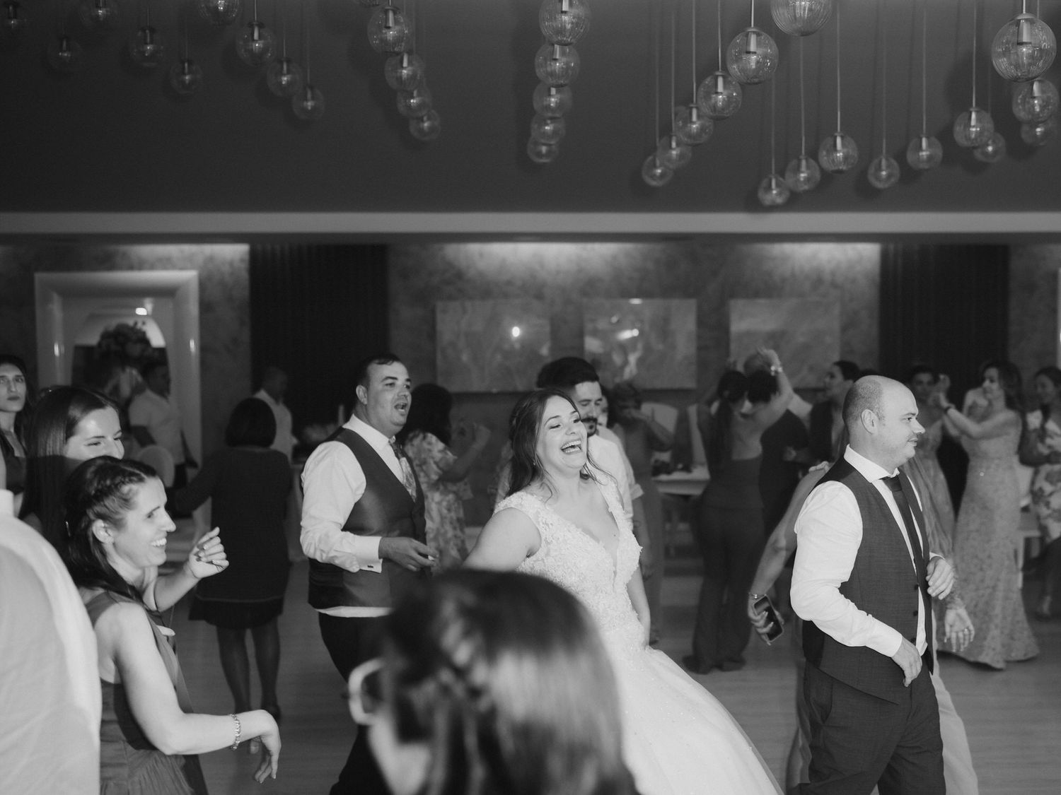 A black and white photo shows couples dancing at an elegant event with hanging lights in the background.