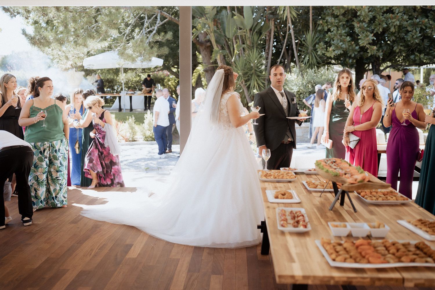 Wedding guests gather at a beautifully decorated outdoor reception with wooden tables and floral arrangements.