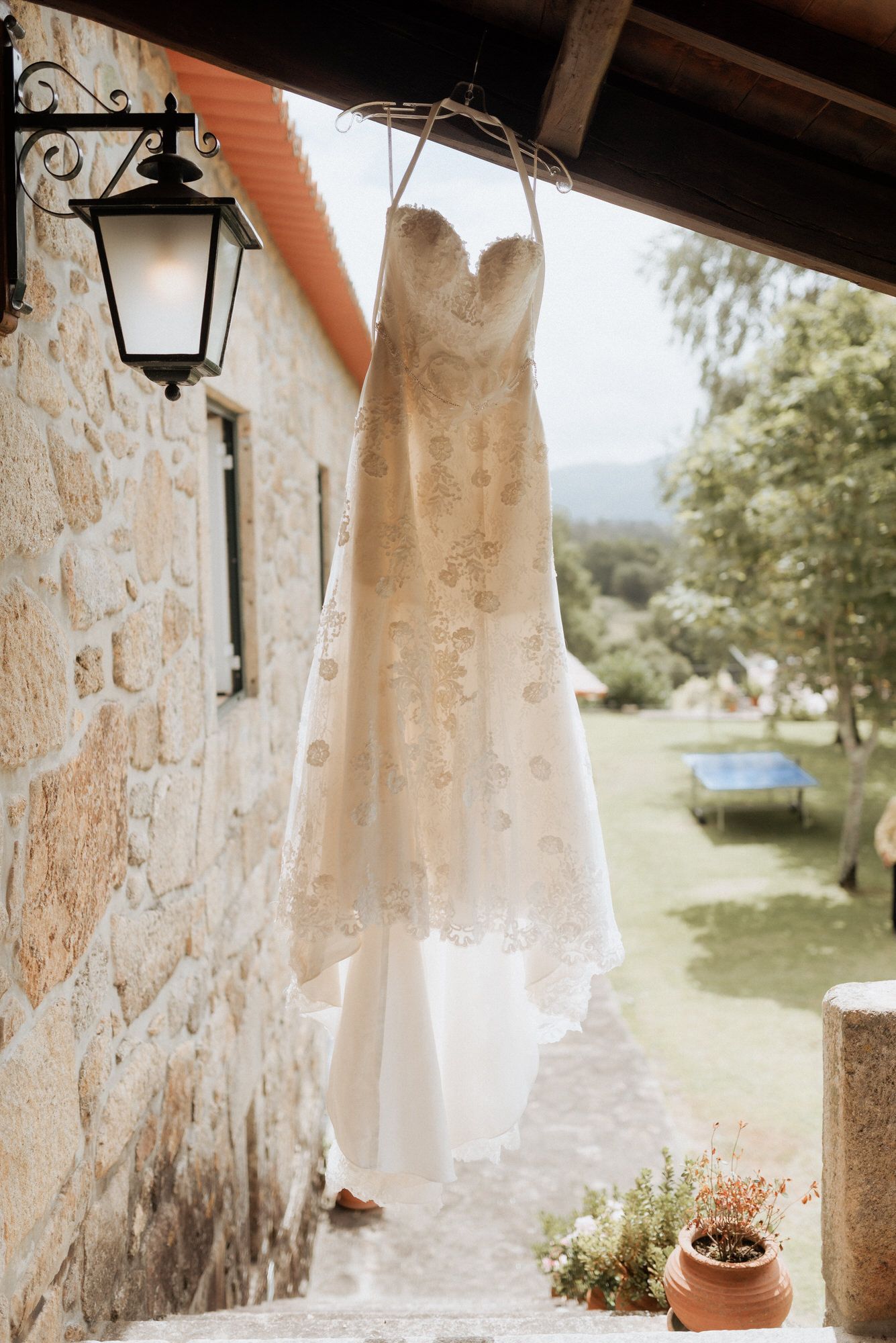 A delicate lace wedding dress hangs from stone wall beside decorative lantern with garden view in background.
