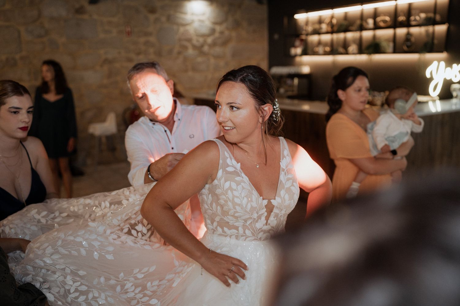 A bride in an elegant lace wedding dress sits at a reception table in a rustic stone-walled venue.