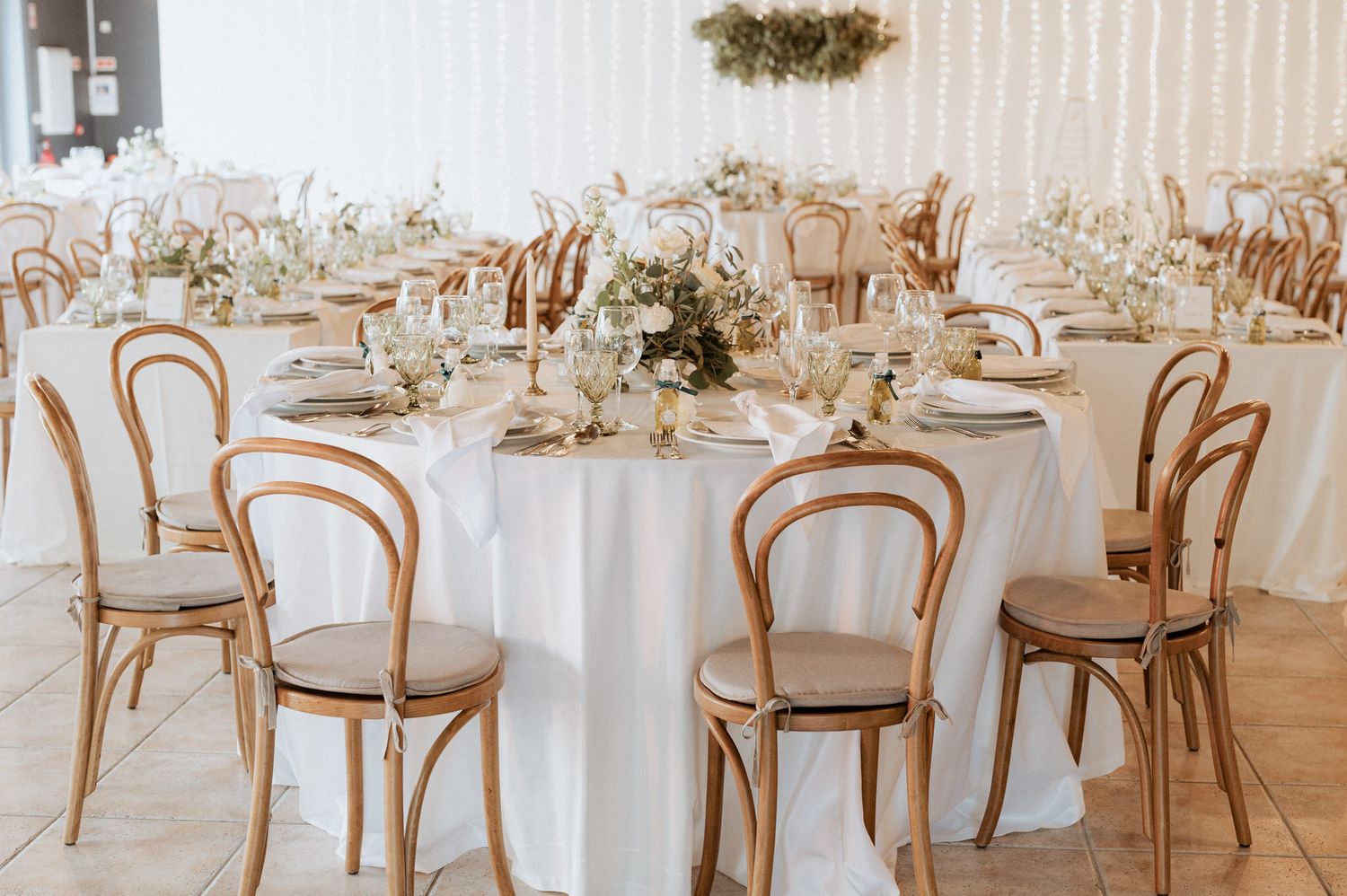 Elegant wedding reception tables with round white tablecloths, wooden bentwood chairs and delicate floral centerpieces.