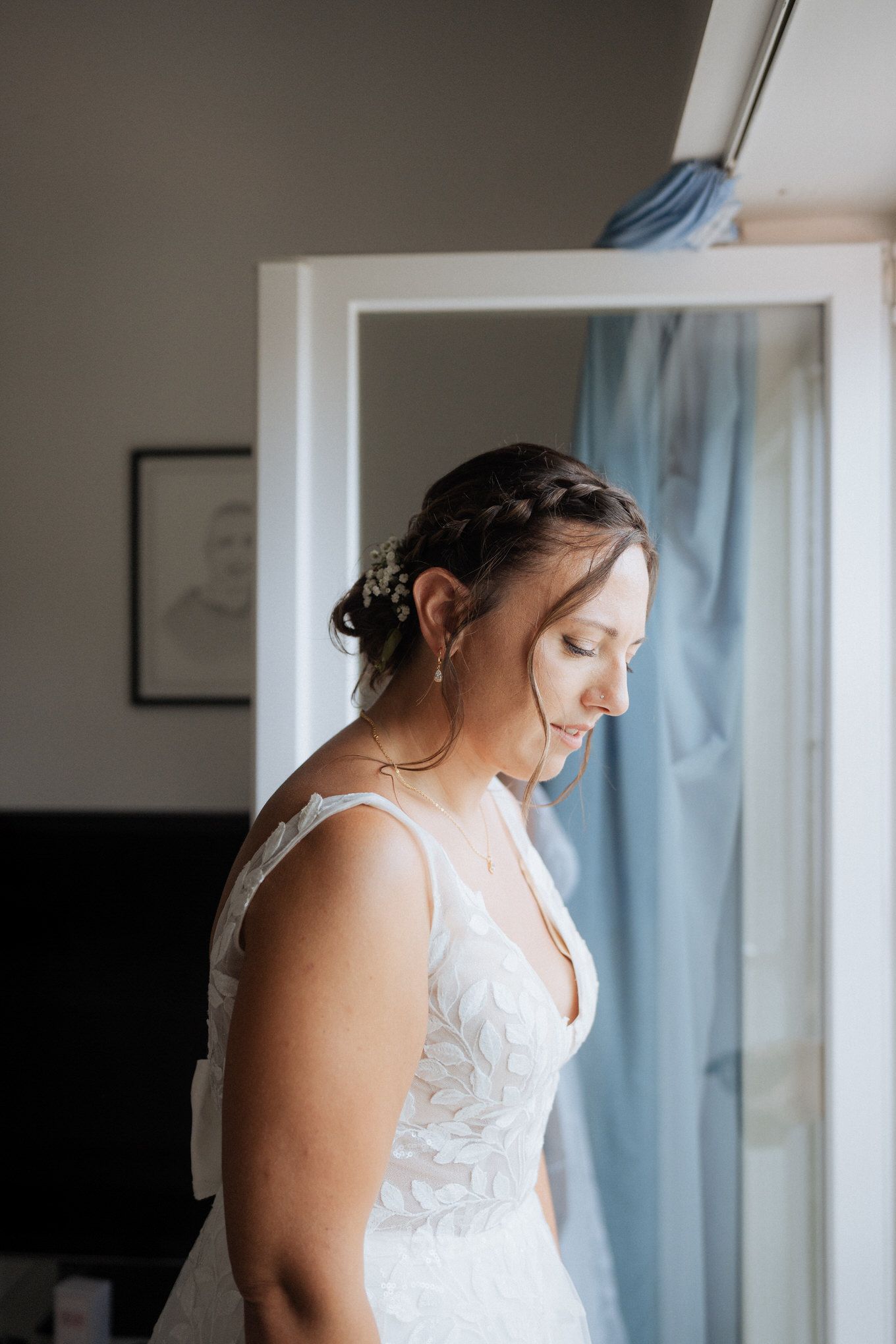 A bride in a white lace wedding gown stands in soft natural light beside a window with blue curtains.