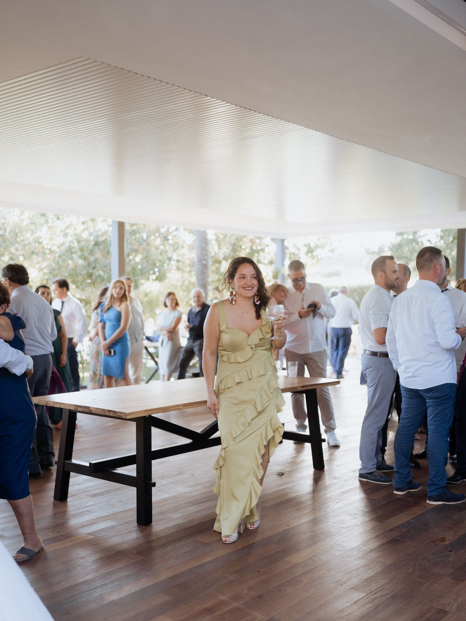 People gather at an indoor event space with hardwood floors and white ceiling as a woman in a gold dress walks past tables.