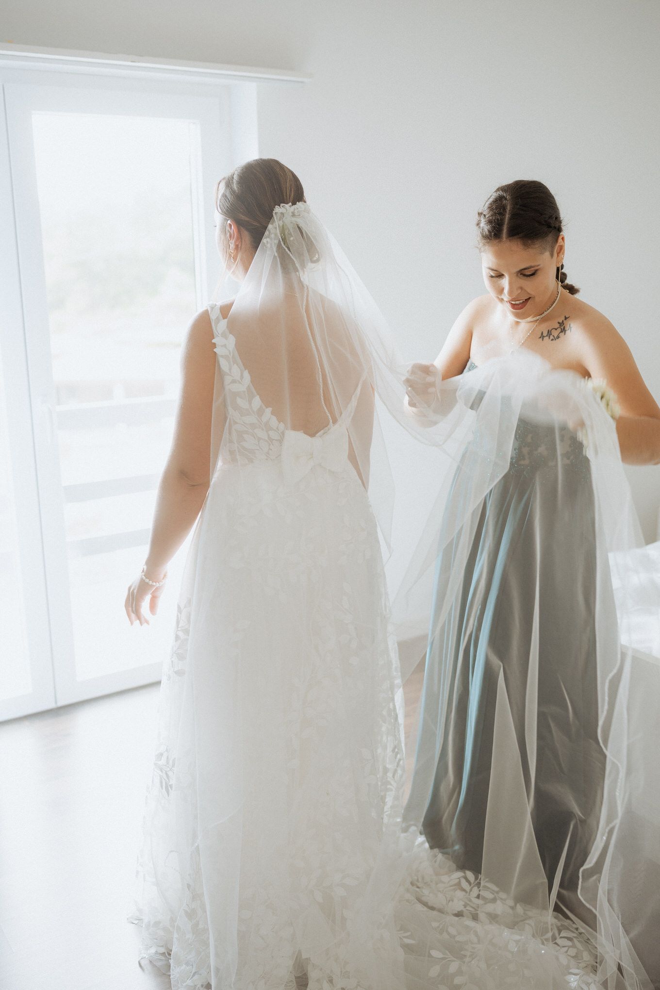 Two women in a bright room on a wedding day, with one in a white lace wedding gown and long veil.