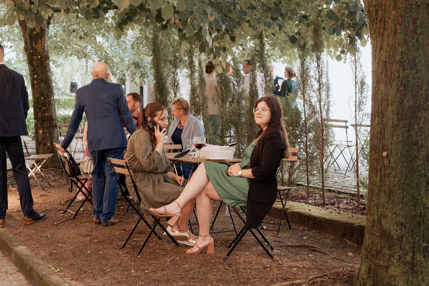 People seated at tables having a casual outdoor gathering under a canopy of trees in natural lighting.