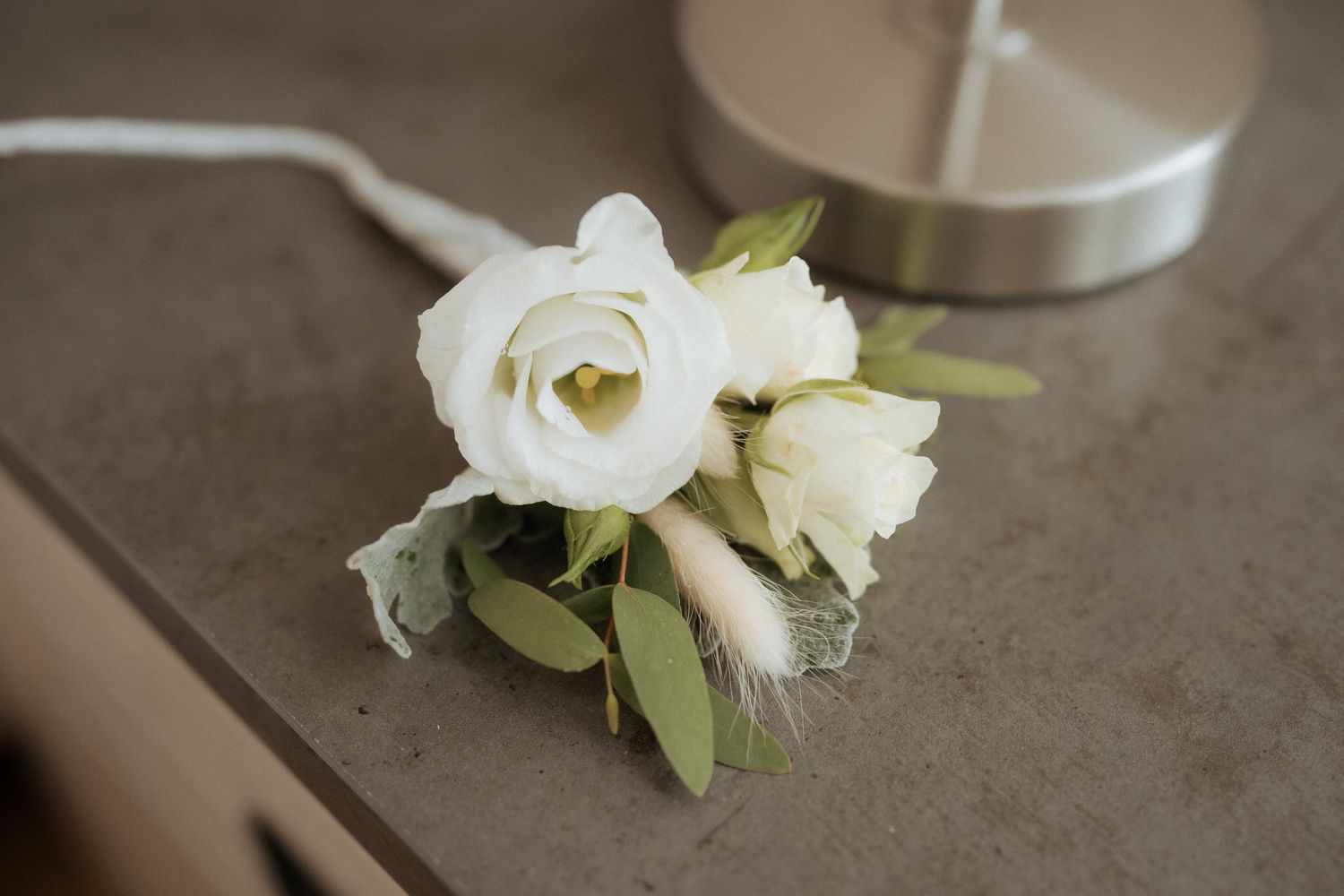 Small bouquet of white lisianthus flowers with green leaves lying on a concrete gray surface near a silver lamp base.