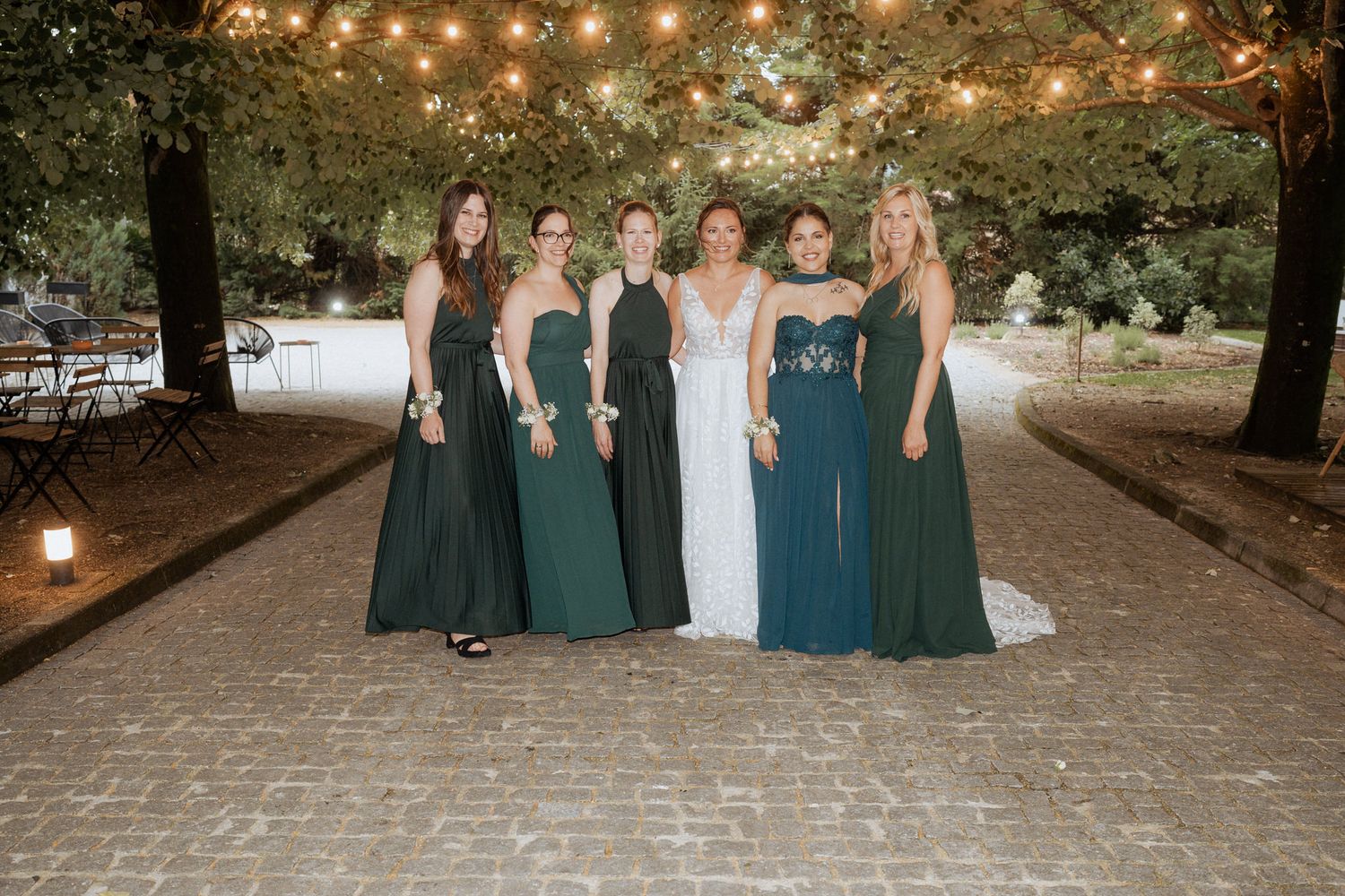 A group in formal evening gowns stand together on a brick path under string lights at an outdoor event.