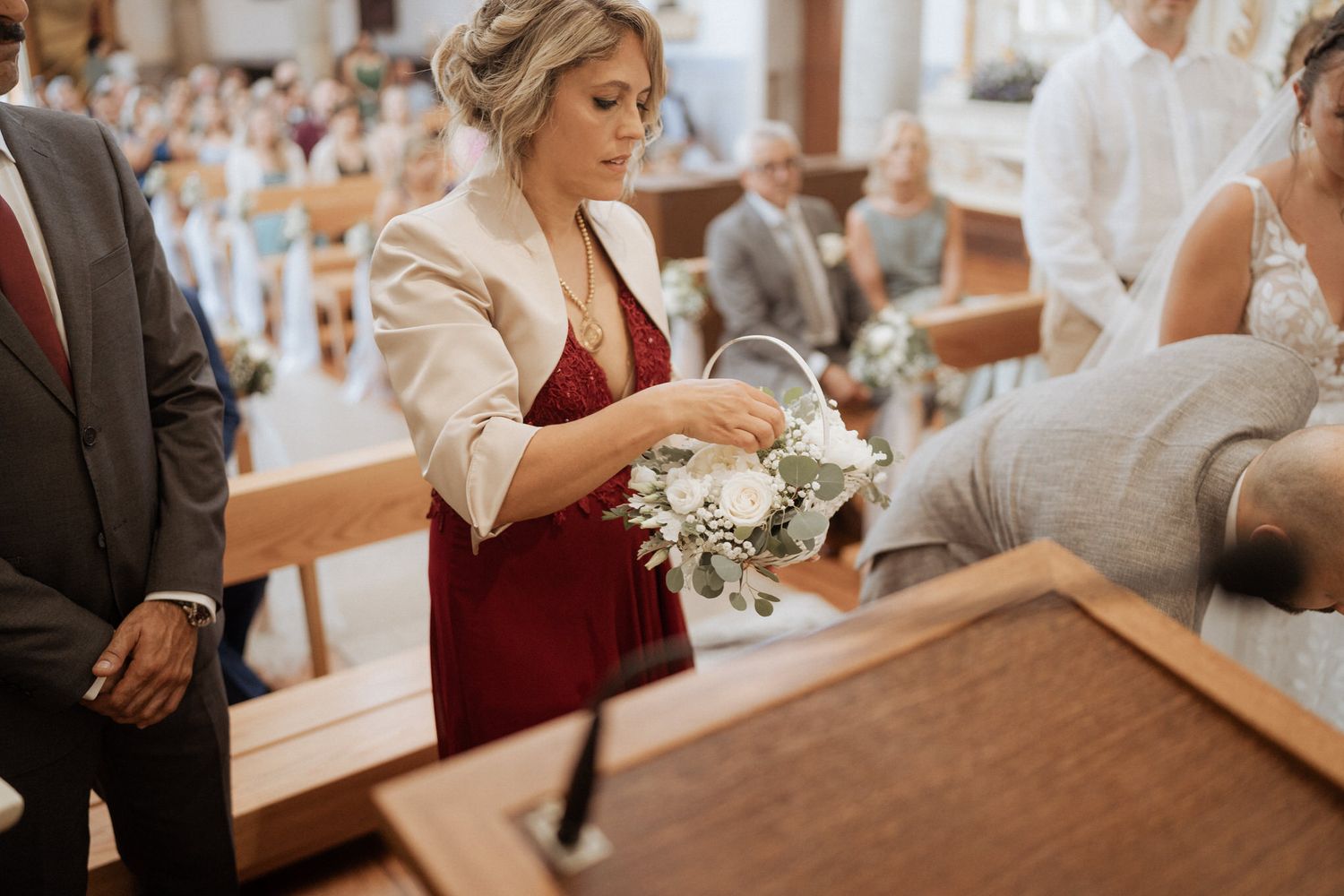 Someone wearing a red dress and cream blazer holds a delicate bouquet during a formal church ceremony.