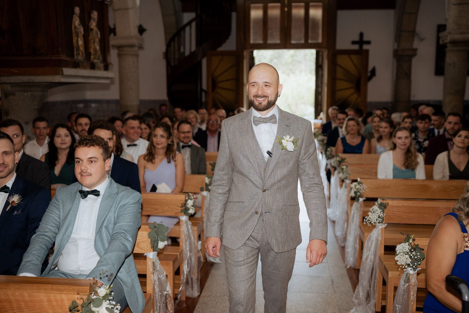 Wedding ceremony in a church with guests seated in wooden pews decorated with floral arrangements.