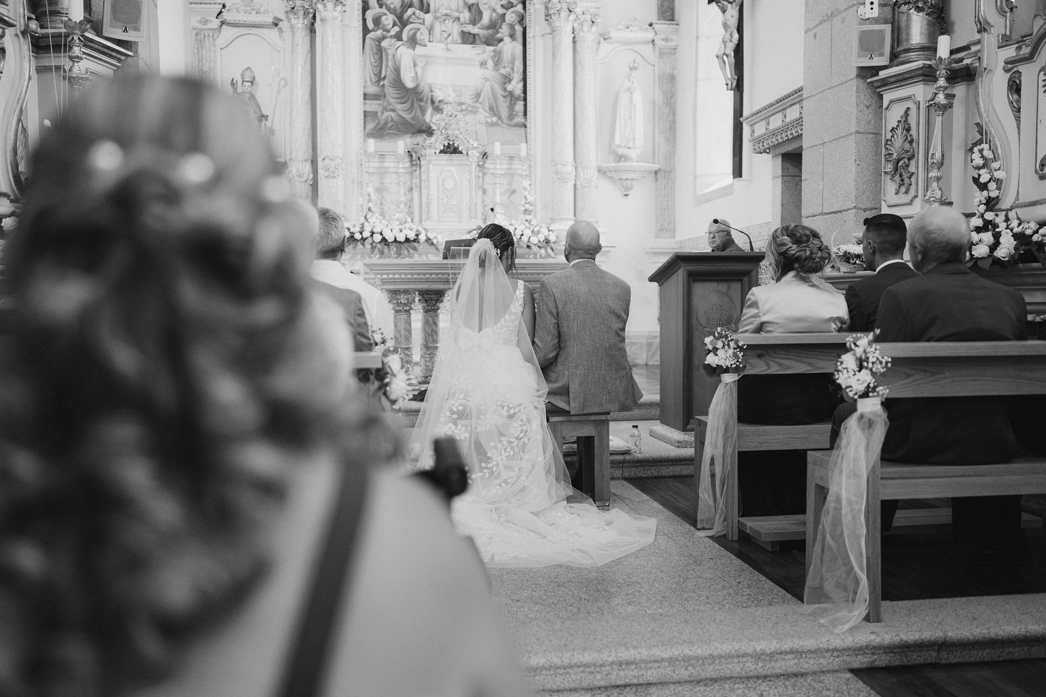 Black and white photo of a traditional church wedding ceremony showing a long white bridal gown from behind in the pews.