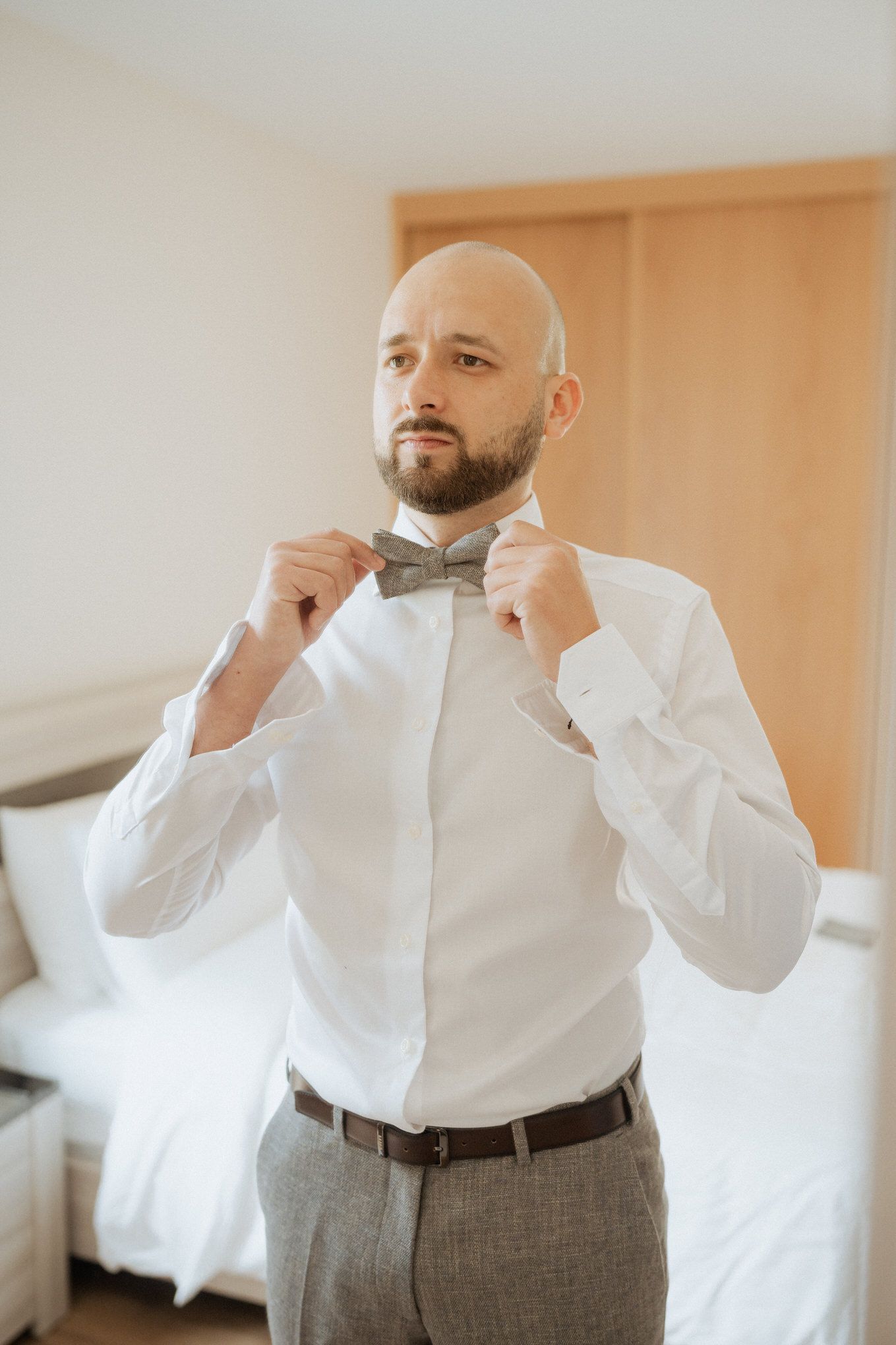 A groom adjusts his bow tie while getting ready in a room with natural lighting and casual elegance.