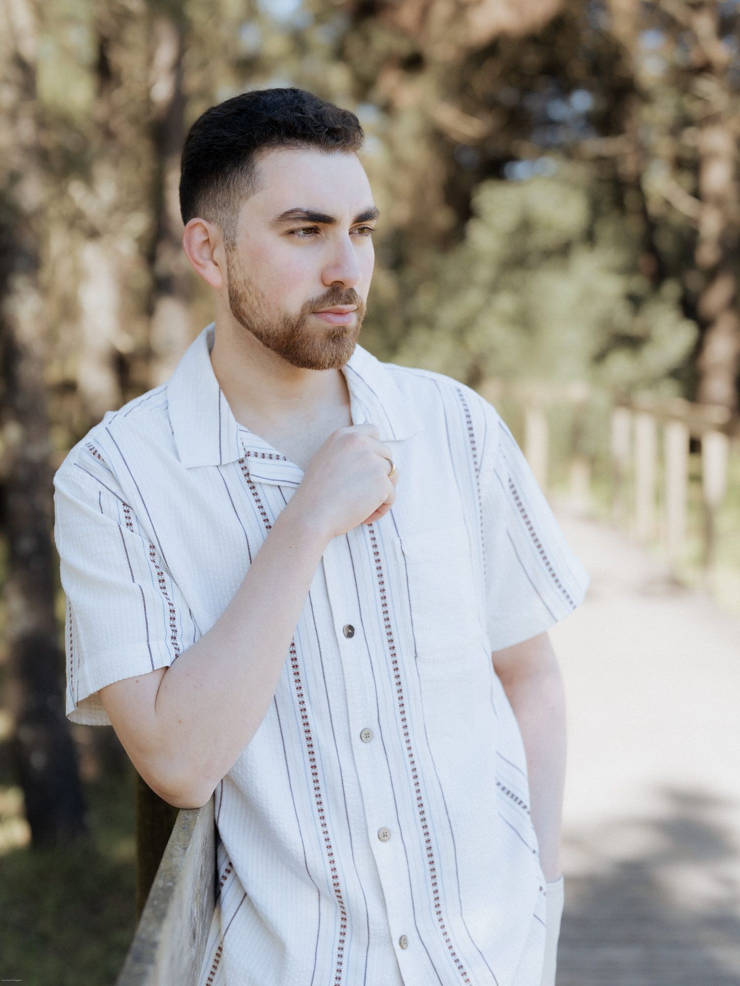 Person in a white patterned short-sleeve button-up shirt posed thoughtfully outdoors on a sunny path.